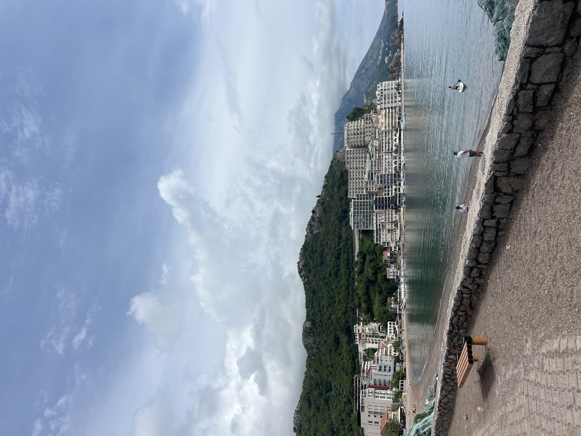 The image depicts a coastal scene in Rafailovići, Montenegro. The view includes buildings on a hillside, a beach, and the Adriatic Sea. People are visible on the beach, some standing near the water's edge and one person paddleboarding further out in the sea. A stone barrier separates the beach from a paved area with a bench. The sky is cloudy.