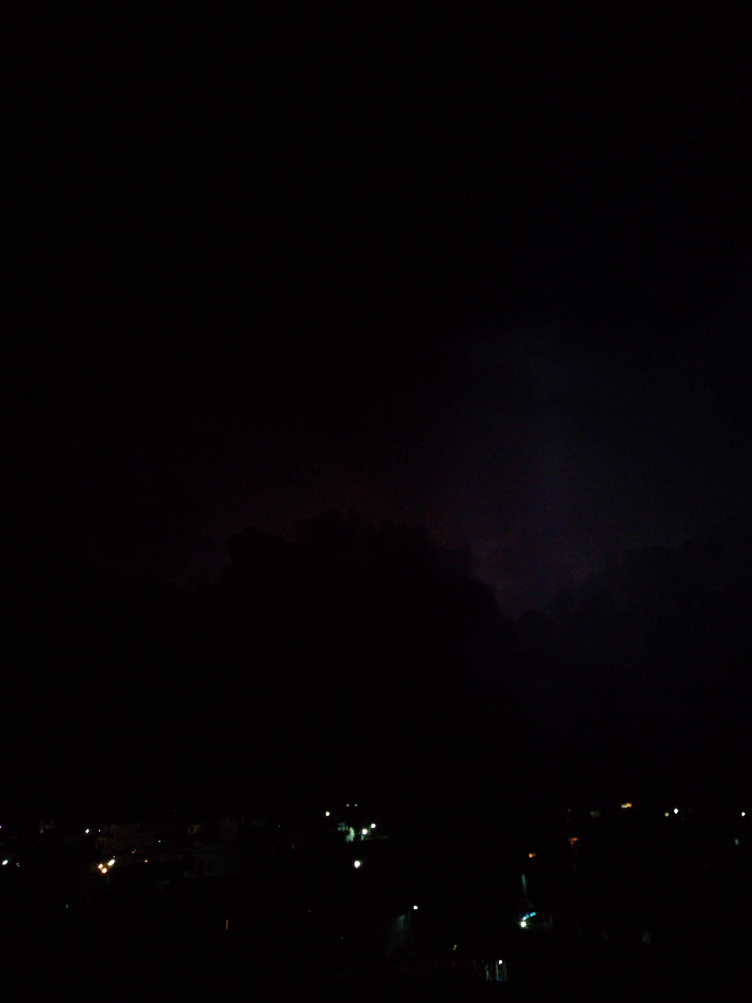 The image depicts a night scene in Phú Quý Special Administrative Region, Vietnam. In the foreground, indistinct city lights twinkle faintly across a dark urban landscape. Above, heavy, dark clouds dominate the sky, punctuated by faint, ominous tinges of purple and red hinting at a possible storm or other atmospheric disturbance. The overall impression is one of darkness and subdued activity in the city below.