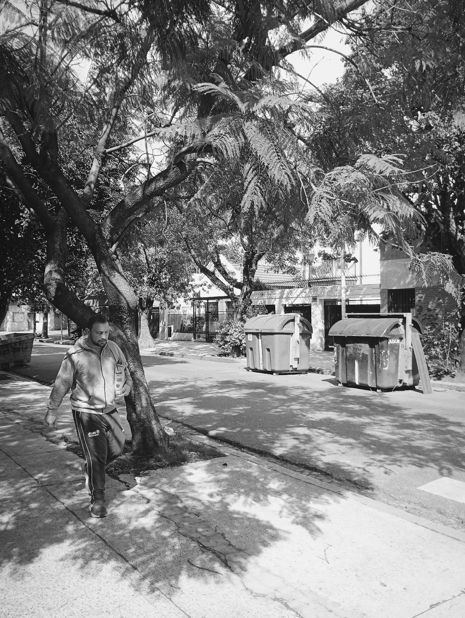 A man walks along a sidewalk in a Buenos Aires neighborhood. He wears a hooded sweatshirt and athletic pants and carries an object in his left hand. To his right, on the street, are two large metal trash containers. A large tree overhangs the sidewalk, casting shadows. Across the street, a house is visible behind a metal fence. The scene is in black and white. The location is identifiable due to the architectural styles and the types of trash containers typically found in residential areas of Buenos Aires.