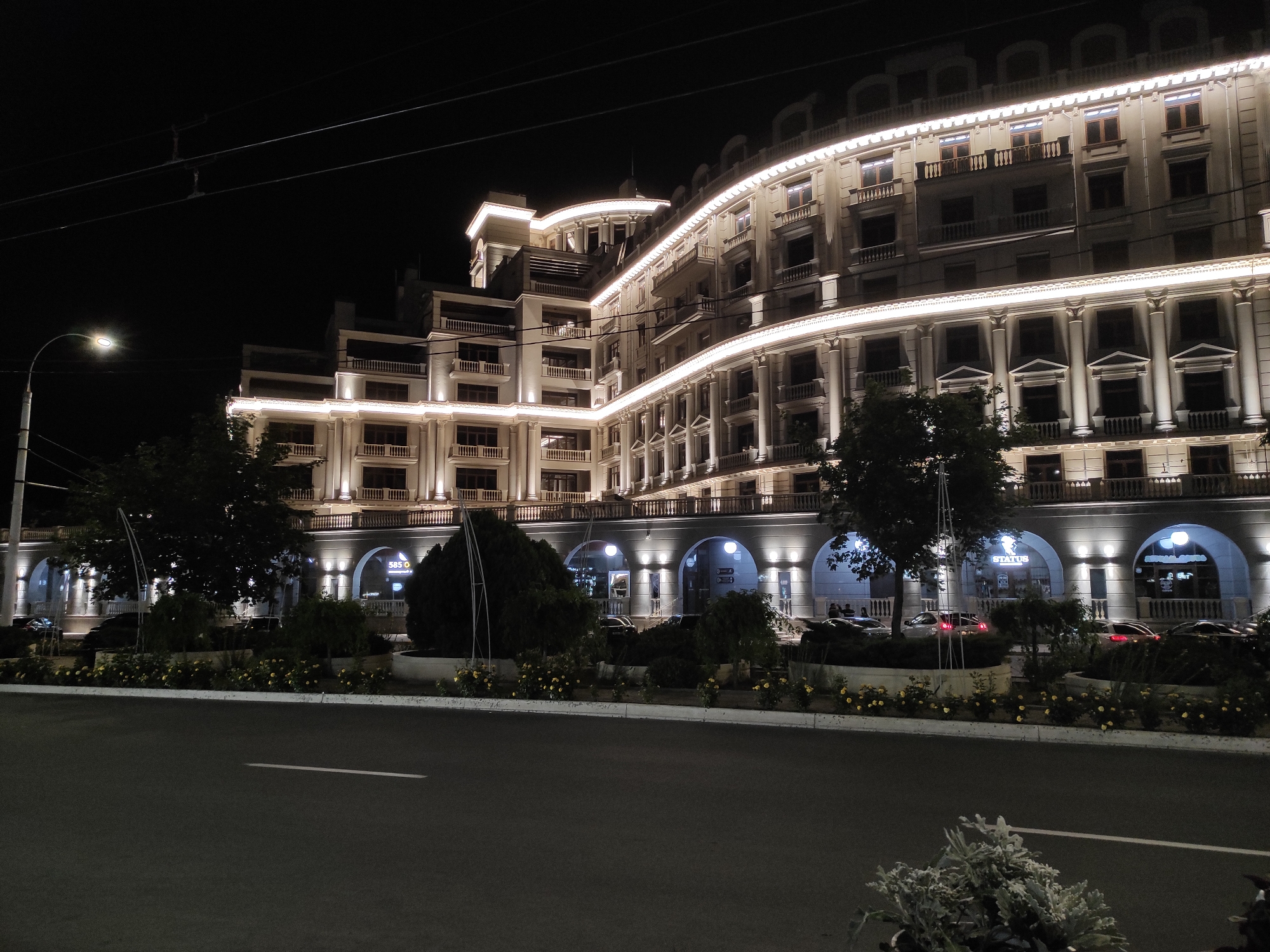 The image depicts a large, ornate building at night in an urban environment. The building is multi-storied with a series of arched entryways at ground level. The facade is illuminated with strings of lights and individual spotlights, emphasizing its architectural features, including rows of windows, columns, and balconies. A road runs along the front of the building, separated by a landscaped median with various plants and small trees. Several cars are parked along the roadside. A lamppost is visible on the left side of the image. The sky is dark, suggesting it is nighttime. The location is Unknown, Unknown.