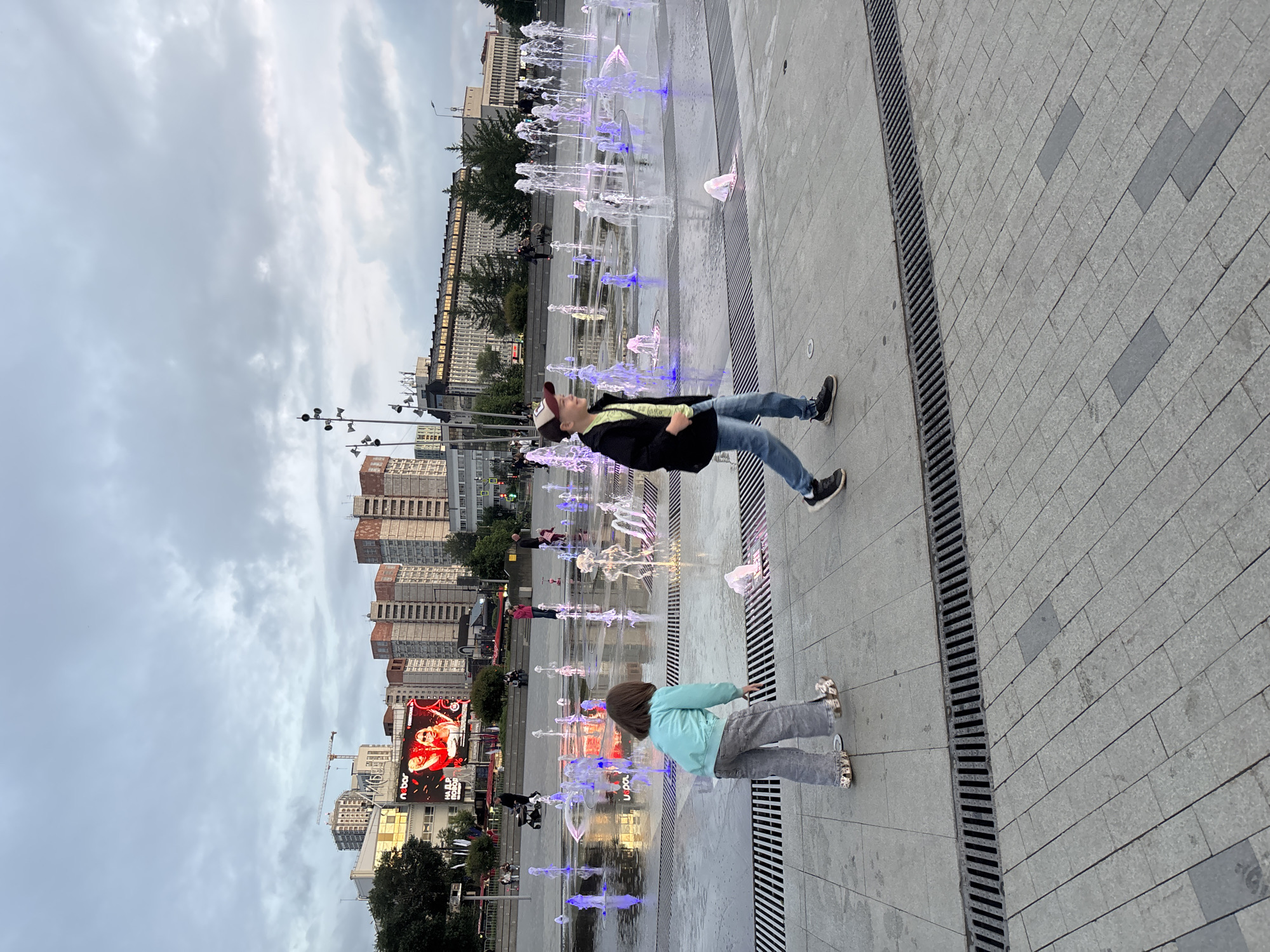 This image depicts a public square in Perm, Russia, identified as the square in front of the Perm Academic Theatre-Theatre. The scene is illuminated under an overcast sky with grey clouds.
In the foreground on the left, a young female, approximately 5-7 years old, with brown hair, wears a light blue jacket and grey pants, standing near the interactive fountain. Her footwear appears to be roller skates or shoes with integrated wheels. To her right, a young male, approximately 8-10 years old, walks towards the left, smiling. He wears a dark jacket, a light-colored t-shirt, blue jeans, and a baseball cap.
The ground is paved with light grey rectangular and square stone pavers, with dark metal grates running along the edges of the fountain area for drainage. The fountain itself is a ground-level installation featuring multiple water jets, illuminated in shades of blue, purple, yellow, and pink, creating reflections on the wet pavement.
In the midground, several people are present, including an individual pushing a baby stroller. The background features various urban buildings. On the left, a large building, identified as the Perm Academic Theatre-Theatre, has a prominent illuminated LED screen displaying an advertisement for "N bar" with an image of two women and additional Cyrillic text. To the right and in the further background, multiple multi-story buildings are visible, including residential blocks with light facades and reddish-brown upper elements, and other structures with darker roofs. A construction crane is identifiable above the buildings in the distant left. Multi-headed streetlights are positioned on the left side of the square.
