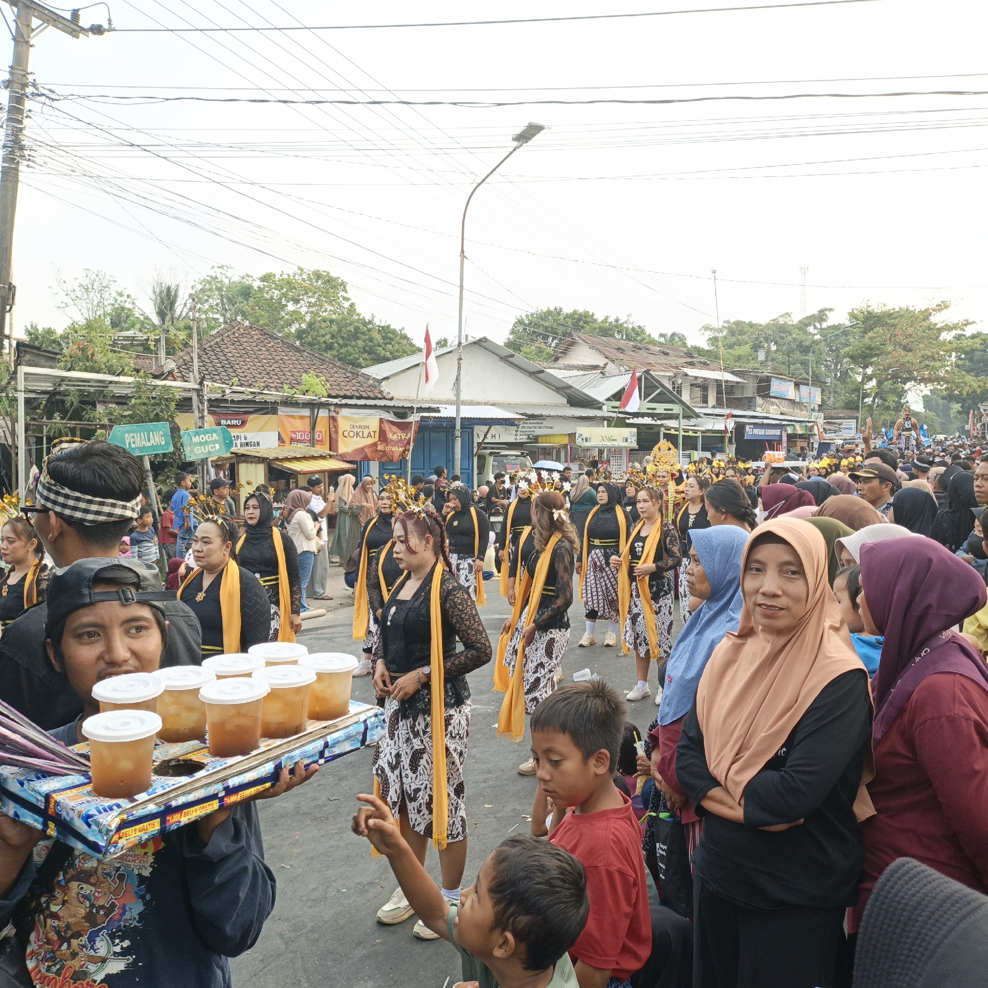This is a medium shot, eye-level, outdoor photograph taken during a daytime event in Randudongkal, Indonesia. The foreground is dominated by a man in a dark blue shirt, wearing a black baseball cap, holding a tray with seven cups of iced tea. The tray is resting on a box wrapped in colorful patterned paper. Behind him, several people are visible, including two young boys, one in a red t-shirt pointing at something off-camera, and another in a green t-shirt.
In the mid-ground and background, a procession is taking place. Several women in traditional black and white attire with bright yellow sashes and headpieces are walking. Other attendees, dressed in various casual clothing and headscarves, line the street. Many are looking towards the procession or the camera. Buildings with tiled roofs and signs, including one that reads "PEMALANG," are visible on either side of the street, suggesting an urban or semi-urban setting. Overhead, a dense network of electrical wires crisscrosses the sky. The lighting is bright, indicating it is likely daytime. The overall atmosphere appears to be festive and communal.
