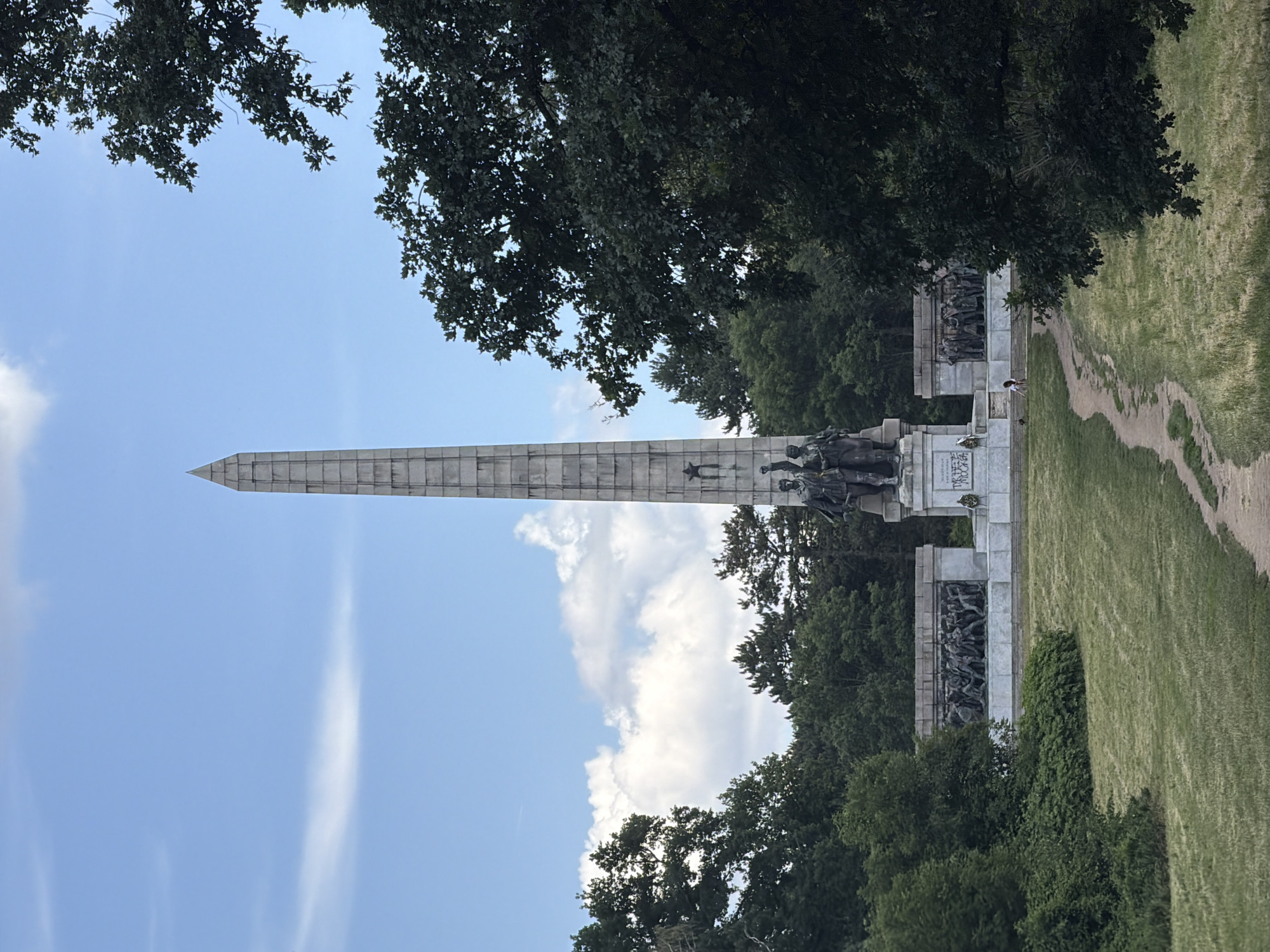 The image depicts a monument on a sunny day, with lush green trees framing the scene. A tall, stone obelisk dominates the left side of the frame, reaching towards a bright blue sky dotted with scattered white clouds. Below the obelisk, a large, ornate monument features intricate sculptures and reliefs depicting groups of figures. These sculptures appear to be made of dark metal, possibly bronze, and are set against a light-colored stone base. Several people are visible near the monument, including a person walking on a path beside a grassy area. The overall setting suggests an outdoor park or memorial site. The time of day appears to be daytime, likely late morning or afternoon, given the bright sunlight casting shadows. The weather is clear and pleasant. The presence of the monument with its historical or commemorative sculptures, along with the verdant surroundings and the clear sky, evokes a sense of solemnity and public remembrance. The location is identified as Sofia, Bulgaria. No visible text is legible in the image.