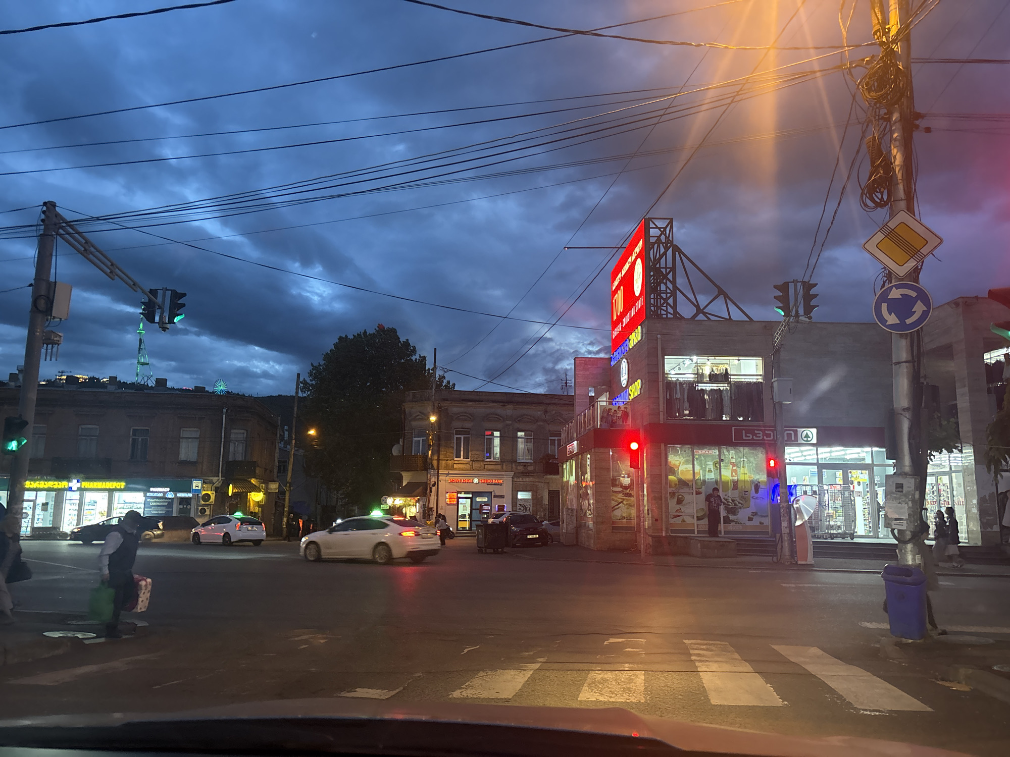 The image captures a street scene in Tbilisi, Georgia, at dusk or twilight. The sky is overcast with heavy, dark clouds. An intersection is visible, with multiple cars and pedestrians present. A pedestrian is crossing the street in the foreground, holding a shopping bag. Buildings flank the intersection, housing various businesses. A pharmacy ("Pharmadepot") is located on the left side of the image. A Spar supermarket can be seen to the right of the frame. Various traffic signals are visible, with one showing green and another red. Overhead power lines stretch across the sky. A roundabout sign is posted on a pole to the right.