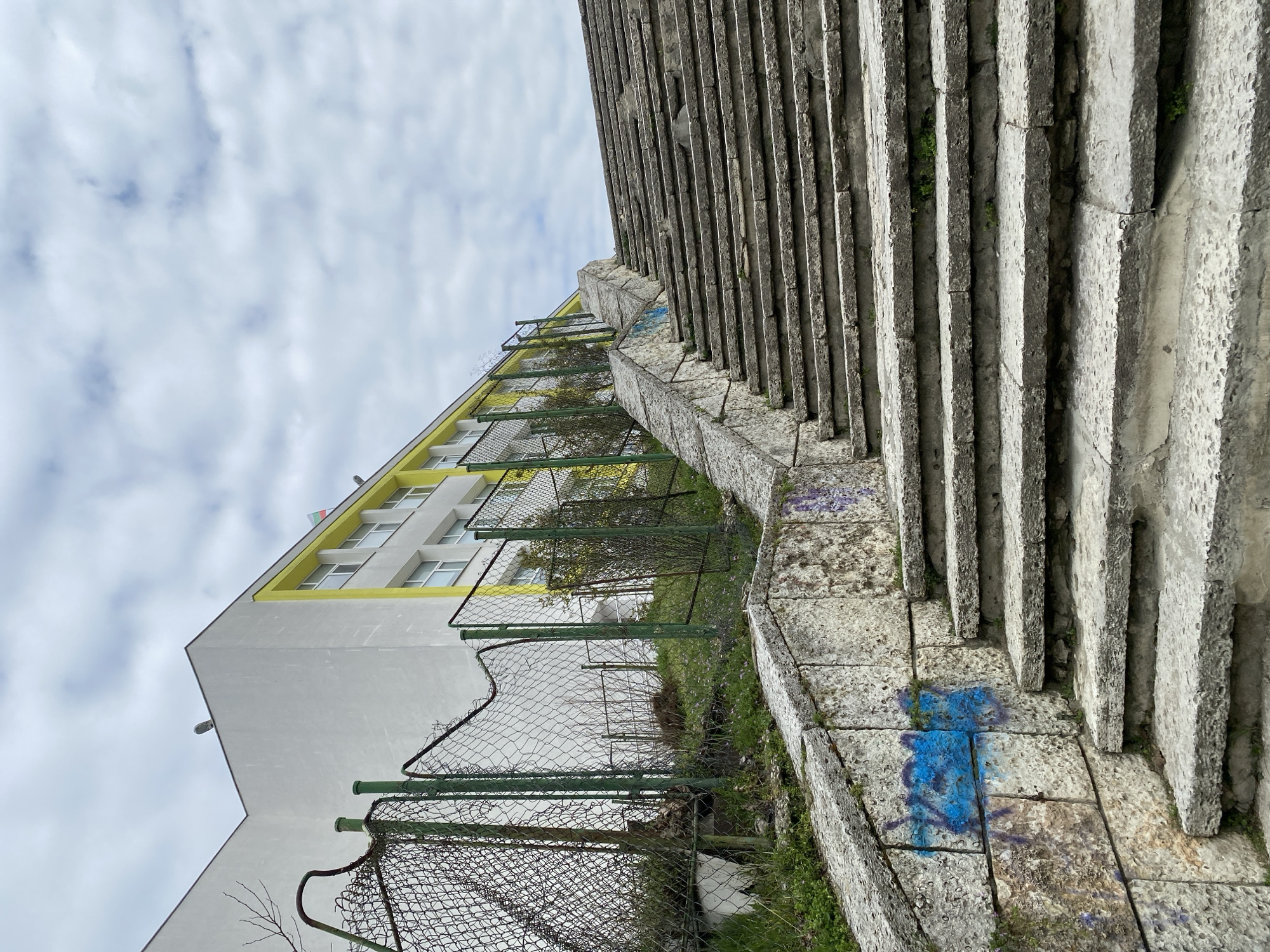 The image shows a set of concrete stairs in Varna, Bulgaria, leading uphill. A building with a white exterior and yellow trim is visible behind a dilapidated green chain-link fence running parallel to the stairs. Graffiti marks the stairs in blue and purple. The sky is overcast with cloudy conditions. The stairs and surrounding area show signs of weathering and disrepair.
