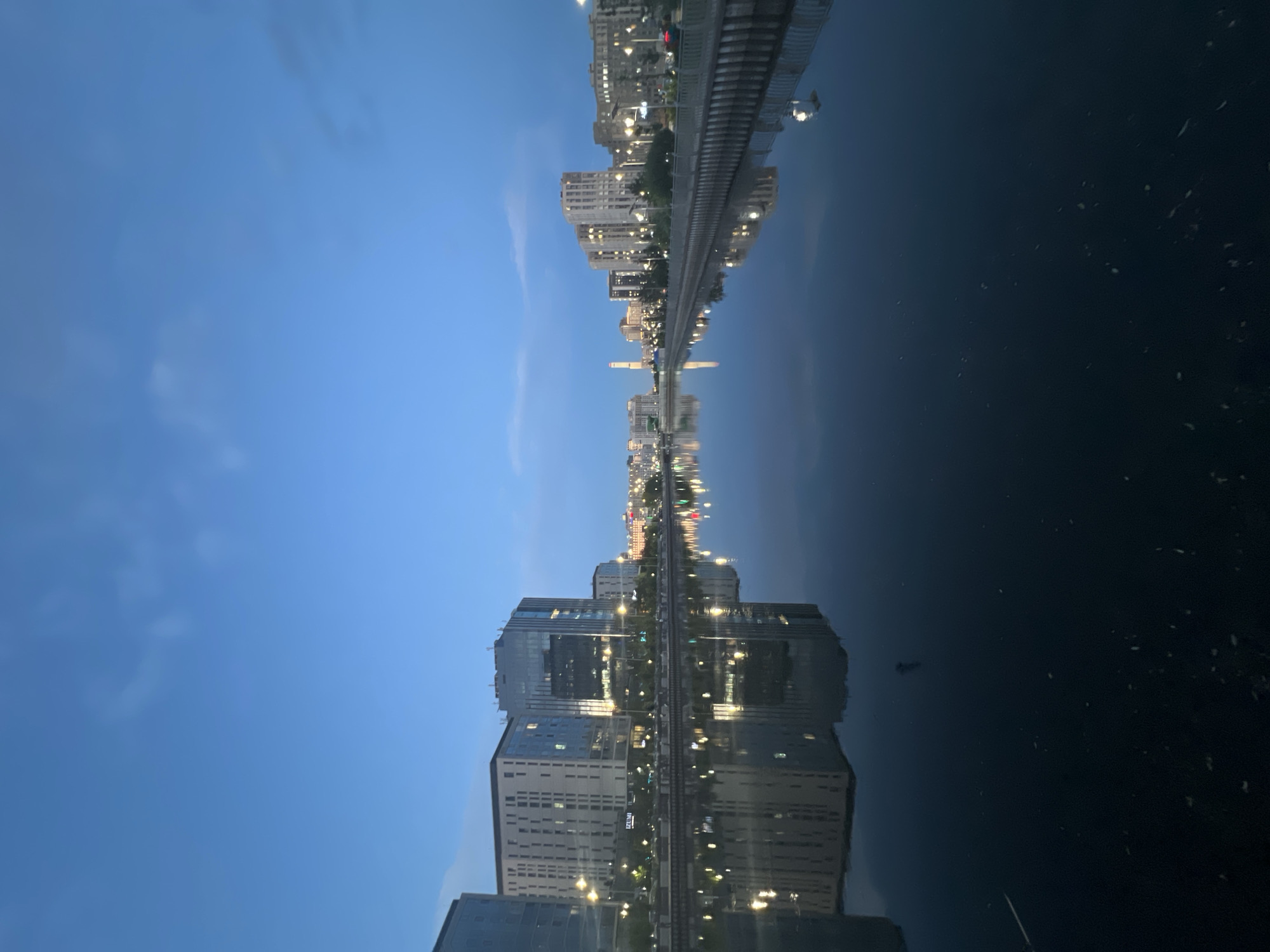This is a nighttime, low-angle, vertical shot of the Ishim River in Astana, Kazakhstan. The sky is a deep blue with faint clouds, and the city lights are reflected on the calm water. On the left bank, modern, multi-story buildings with illuminated windows line the riverfront. On the right bank, similar buildings are visible, as well as streetlights and a walkway. The overall impression is one of urban tranquility at dusk or dawn.