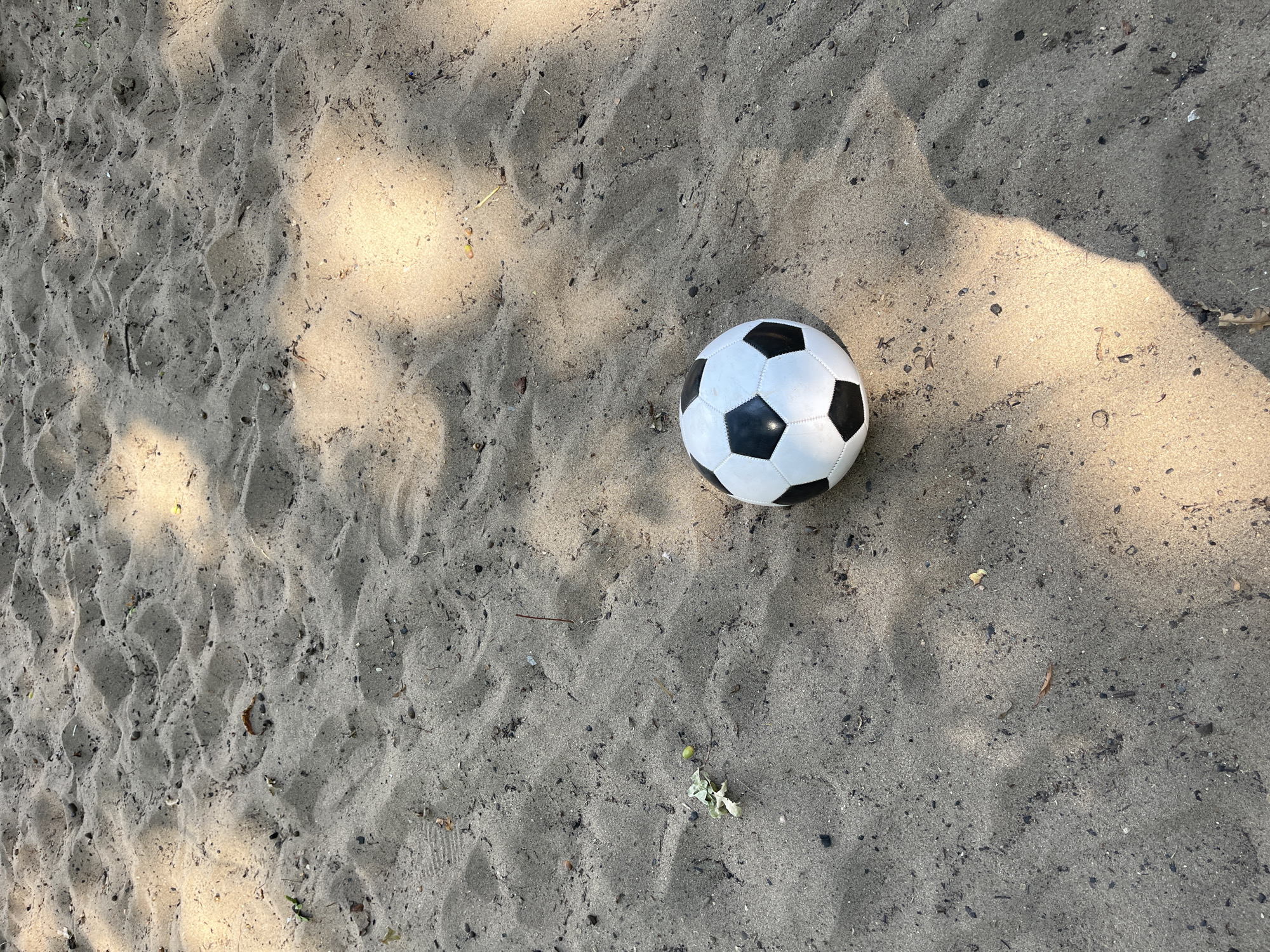 A black and white soccer ball rests on a sandy surface. The sand shows evidence of rippling and small debris such as pebbles and dried leaves. Sunlight filters through what appears to be tree cover, casting dappled shadows across the ground. The scene is set in an outdoor environment, possibly a park or playground, in Saratov, Russia.