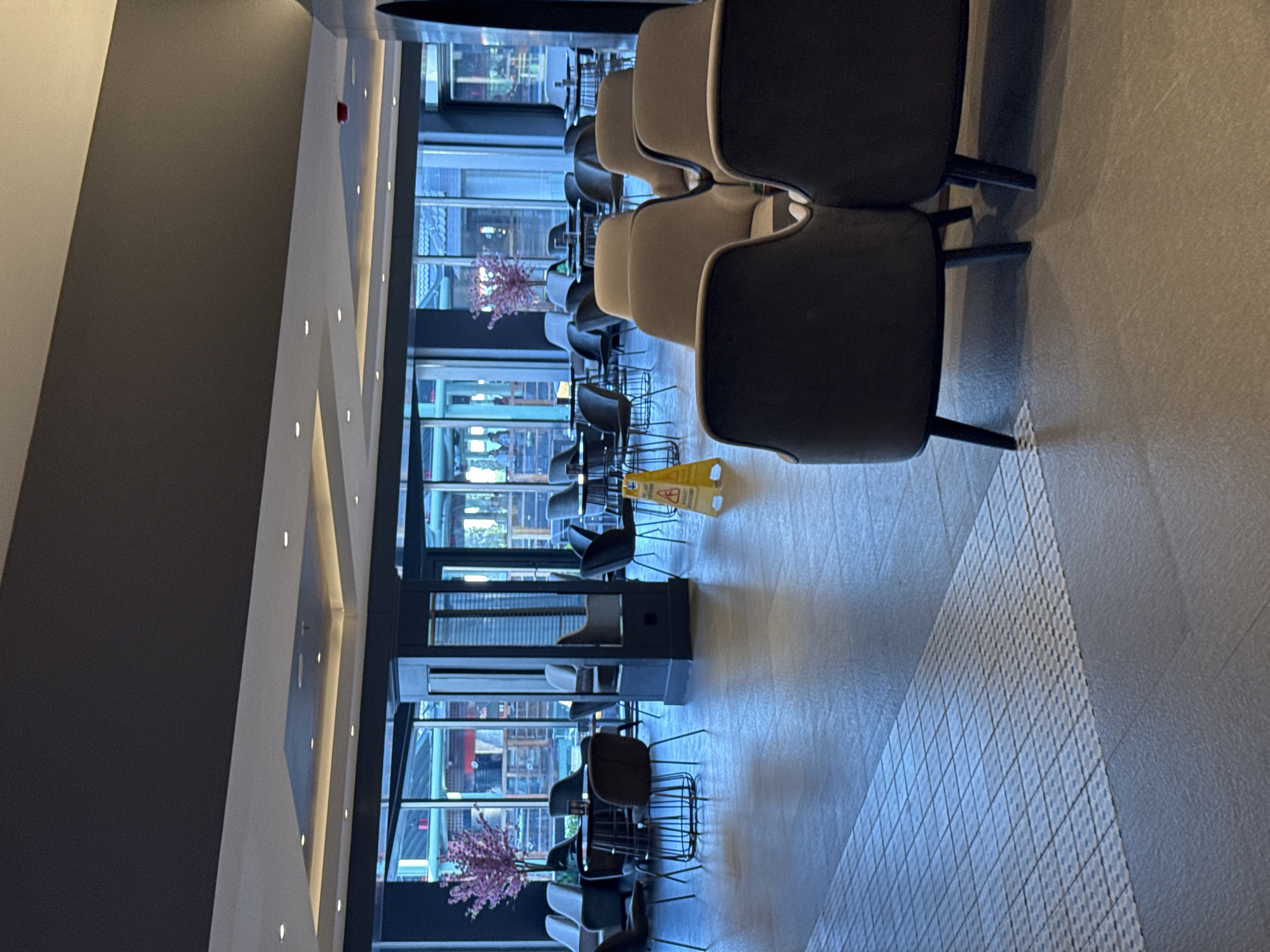 This is a photograph of the interior of a modern cafe or restaurant in Aksu, Turkey. The space is furnished with multiple tables and chairs, with large windows in the background offering a view of an outdoor area with more seating and greenery. The lighting suggests it might be evening or a cloudy day. The flooring is tiled, and there is a caution sign on the floor. There are no people visible in the image, but the arrangement of furniture implies it is a public space meant for dining or relaxation. The decor is contemporary and somewhat minimalist.