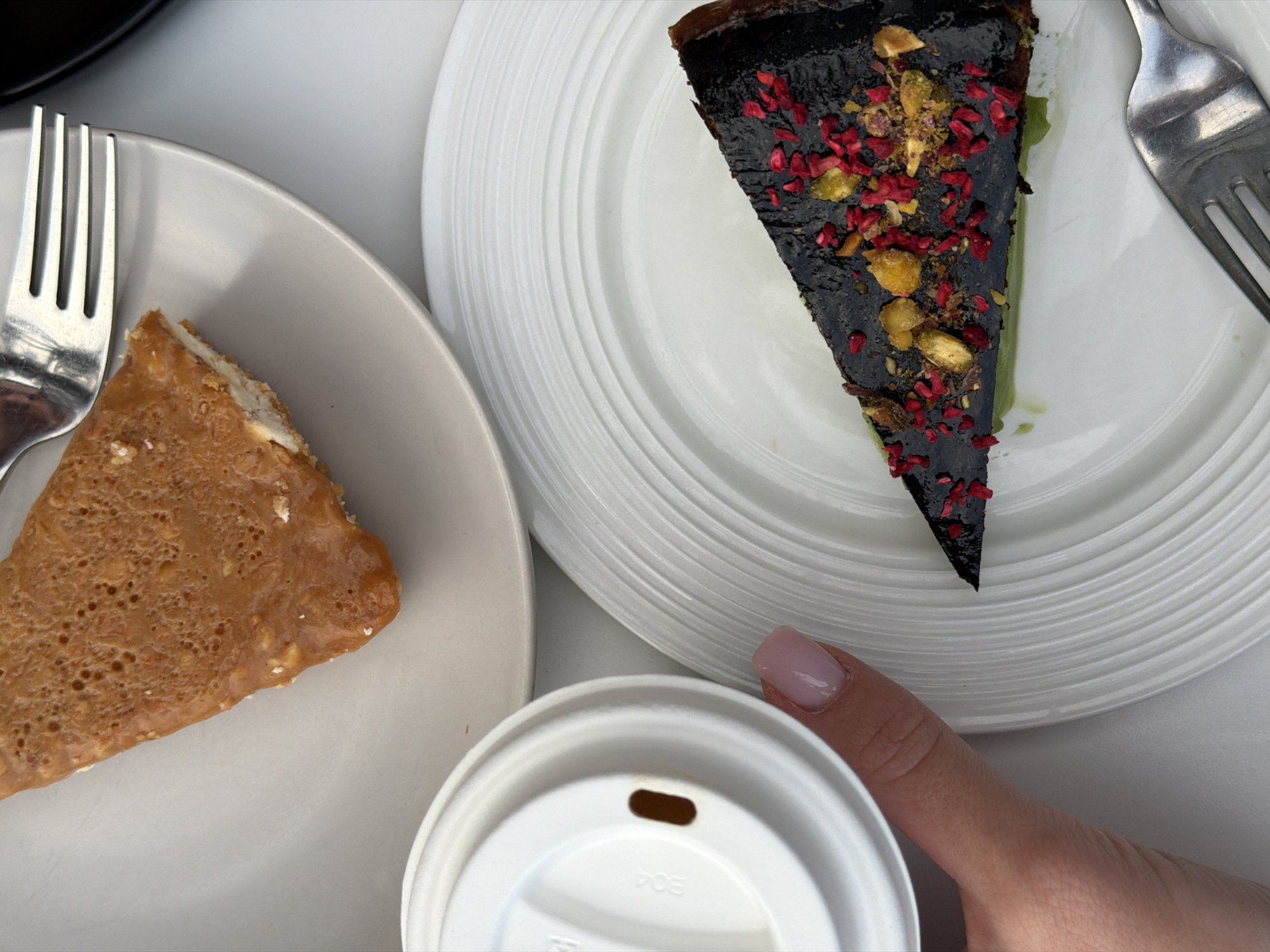 The image presents a still-life composition featuring two plates of dessert and a cup, with a hand partially visible. The scene is likely an overhead shot.
On the left, a triangular slice of what appears to be a caramel or toffee-flavored dessert rests on a pale, slightly textured plate. A silver fork is positioned to the upper left of the dessert slice.
To the right, a slice of dark-colored cake, possibly chocolate, sits on a white plate with concentric ridged circles. The cake is garnished with red and golden-colored toppings. A silver fork is also placed to the upper right of the cake.
A white cup with a lid, bearing text "BO4" is placed in the bottom center. A human hand, with a nail painted in a pale pink color, is partially visible near the cup.
The background consists of a white surface, suggesting a table.
No explicit details are present in the image to confirm the location as Bečići, Montenegro.