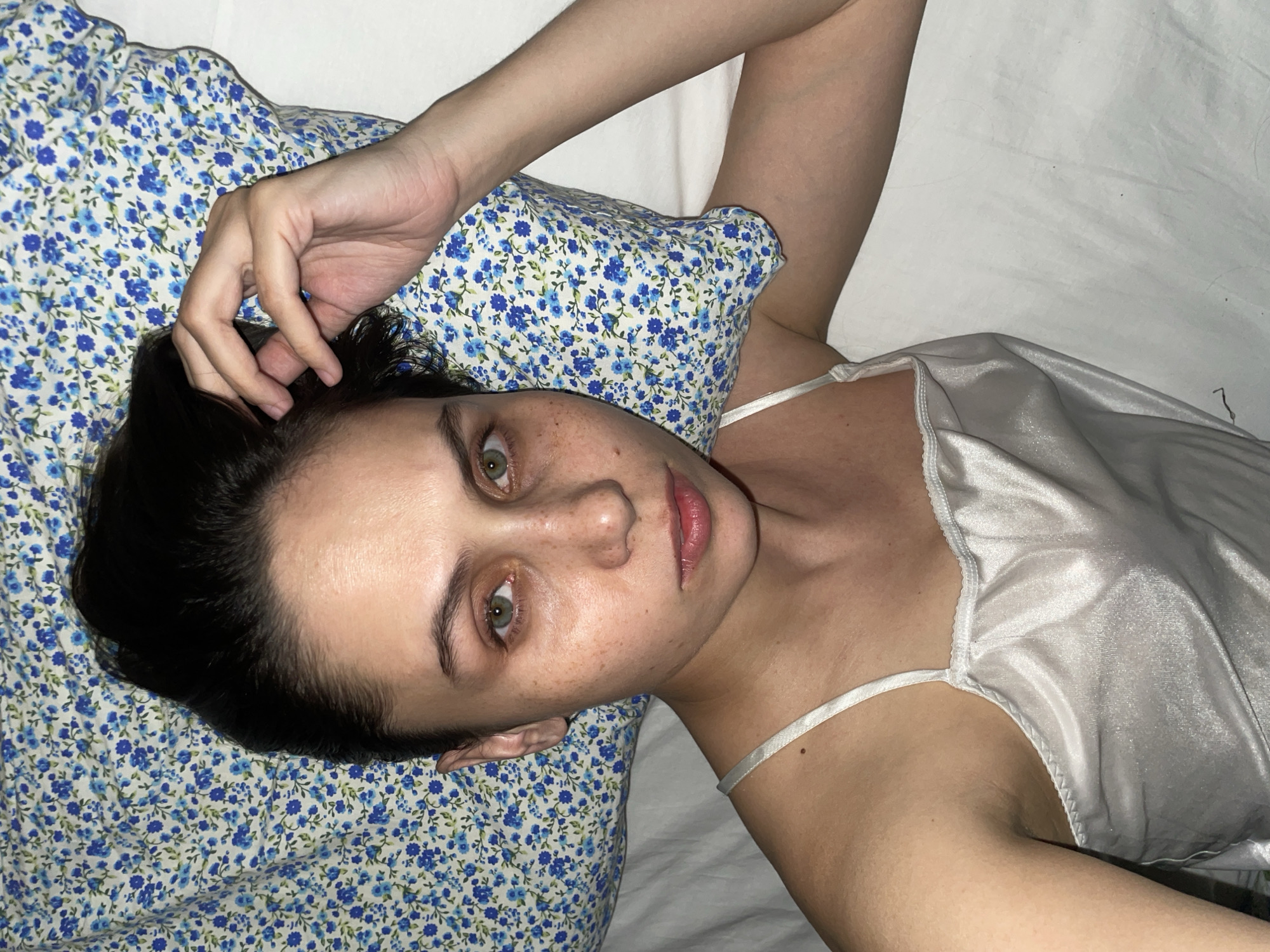 A fair-skinned young woman lies on a bed. Her head rests on a pillow with a small blue floral pattern. She is wearing a light-colored satin camisole. Her hair is dark, and her eyes are green. Her left hand is touching her hair. The background consists of a white bedsheet. There is no evidence to suggest the location is Izhevsk, Russia.