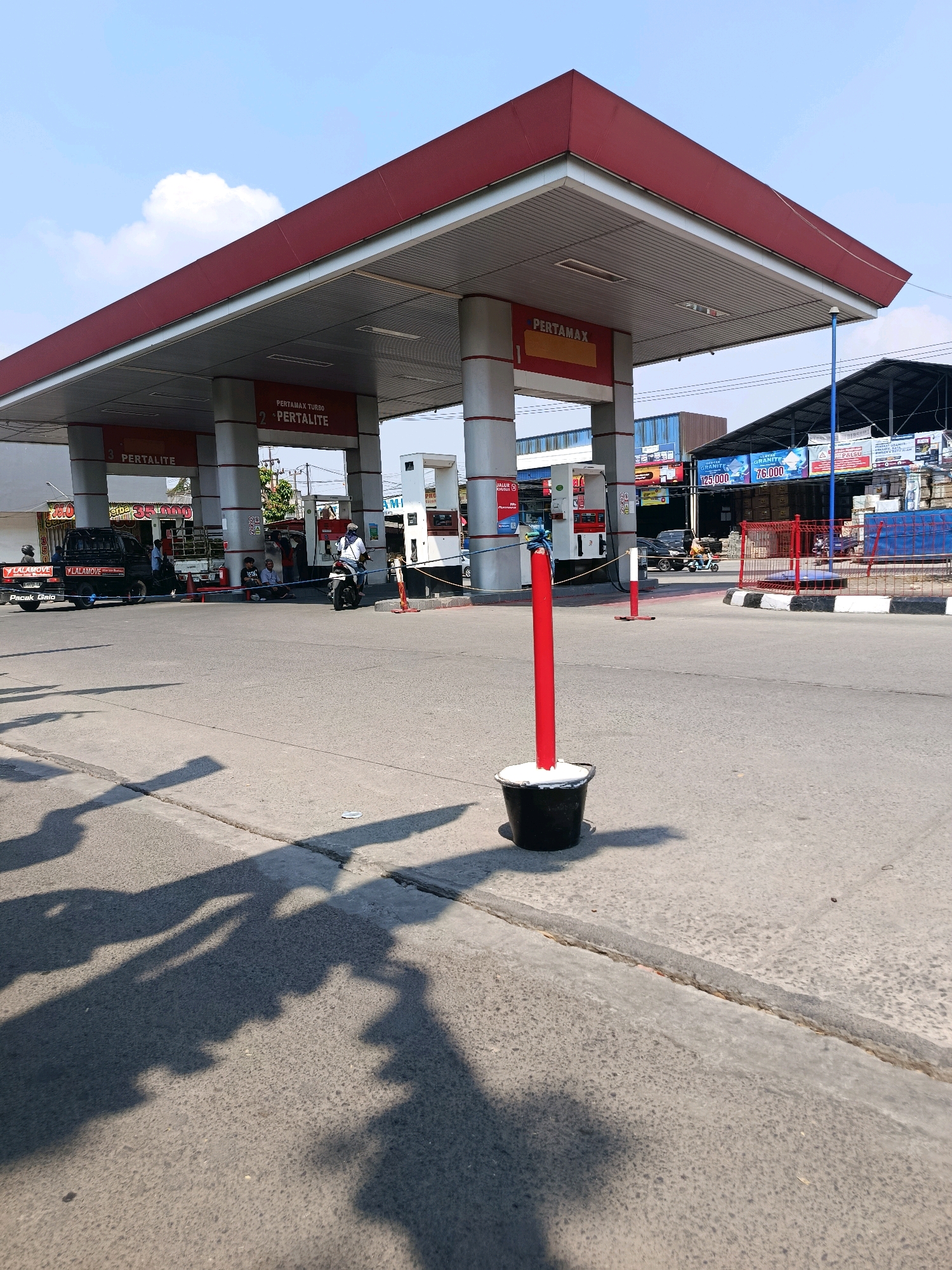 An exterior, ground-level, eye-level, medium shot captures a Pertamina gas station in Babelan, Indonesia, under a clear, blue sky with a few white clouds. The gas station features a red-roofed canopy supported by white pillars, with "PERTAMAX" and "PERTALITE" signs visible. Several gas pumps are positioned under the canopy. In the foreground, a black bucket with white material inside sits on the asphalt, topped by a red pole. The asphalt road shows shadows cast by the gas station canopy.
Several vehicles are present: a black Lalamove delivery truck, a black car, and a white motorcycle. People are visible near the pumps and on the sidewalk. Some are standing, one is sitting on the ground, and another is on the motorcycle. Behind the gas station, a building with a corrugated metal roof and various advertisements is visible. The scene depicts a typical operational day at a gas station, with vehicles and pedestrians engaged in their activities.