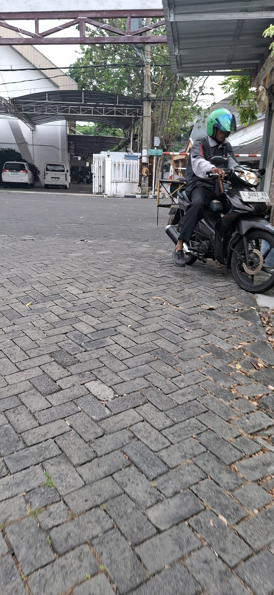 A man in a green helmet and a gray and black jacket is seated on a black motorcycle with a white license plate that reads "L 6002 RB". He is wearing black pants and dark-colored footwear. The motorcycle is parked on a herringbone-patterned brick driveway in front of a white building with large metal gates. A white van and a white car are parked to the left of the building. In the background, trees and power lines are visible. A street sign with green text and a white border is mounted on a pole to the right of the gates. The scene appears to be an outdoor area within a city, likely an entrance to a residential or commercial property. The lighting suggests it is daytime, possibly overcast.