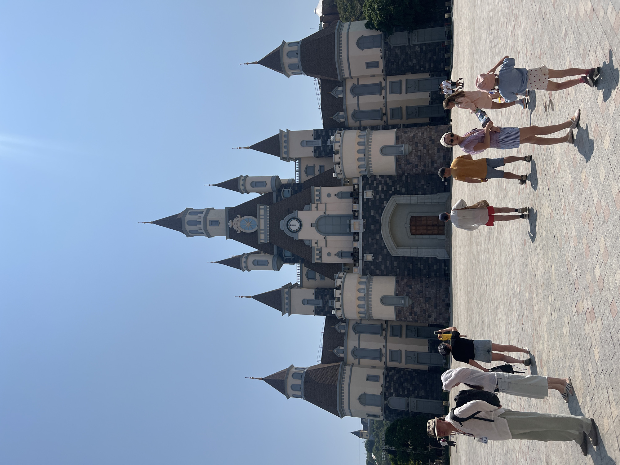 The image depicts a group of people standing in front of a large castle. The castle is multi-layered with multiple towers and spires. It has a predominantly light beige exterior, accented with brown and gray details, and features architectural details such as windows and arches. Two visible clocks are installed on the facade.
The people in the image are standing on a paved surface in the front of the castle. They are dressed in casual attire, including shorts, shirts, and hats. Some people are holding cameras or phones, as if taking pictures of the castle.
Based on the style of the castle, the location can be identified as VinWonders Nha Trang, a theme park on Hòn Tre Island, Nha Trang, Vietnam. The environment is bright, suggesting a sunny day.