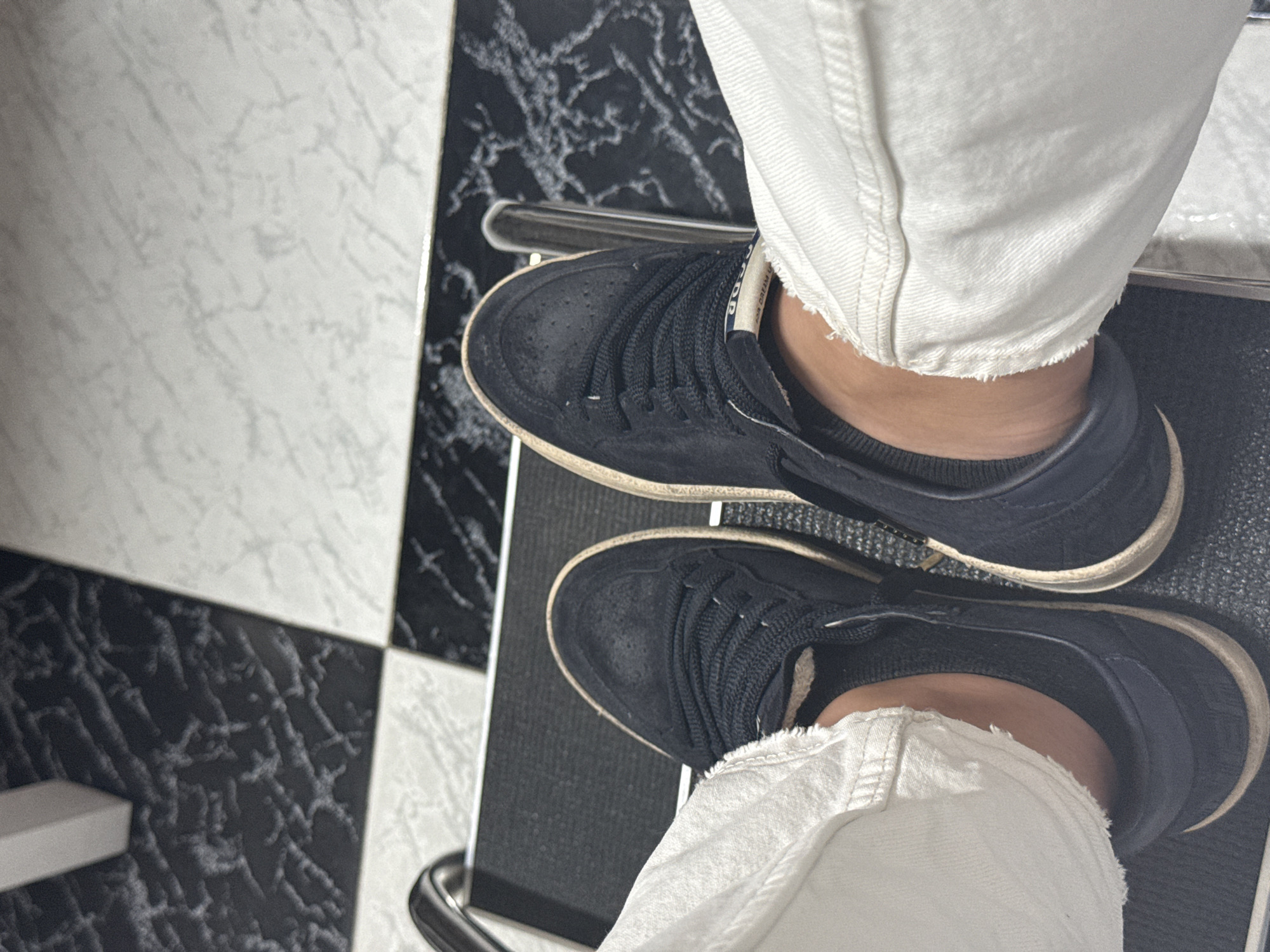 The image captures a close-up, high-angle view of a person's lower legs and feet as they sit, resting their feet on the base of a chair or stool. The scene is indoors, likely in Puebla, Mexico, as per the provided context.
The subject is wearing a pair of dark navy or black sneakers, possibly made of suede, with off-white or cream-colored soles that exhibit a worn or distressed appearance. A white label on the tongue of the right shoe is partially visible, showing text that includes "G.G.D.P" and "FOR DREAM," strongly indicating the brand Golden Goose Deluxe Brand. The sneakers are paired with casual white pants, likely jeans, which have a distinctive frayed or raw hem around the ankles. The person appears to be wearing dark, possibly black, ankle socks.
The feet are positioned on a dark, possibly black, surface that is part of the chair's structure, supported by a shiny, metallic, possibly chrome, frame. The floor features a striking checkerboard pattern of black and white tiles, both of which are patterned with marble-like veining, creating a sophisticated yet graphic background. The overall impression is one of casual style in a distinctive interior setting.