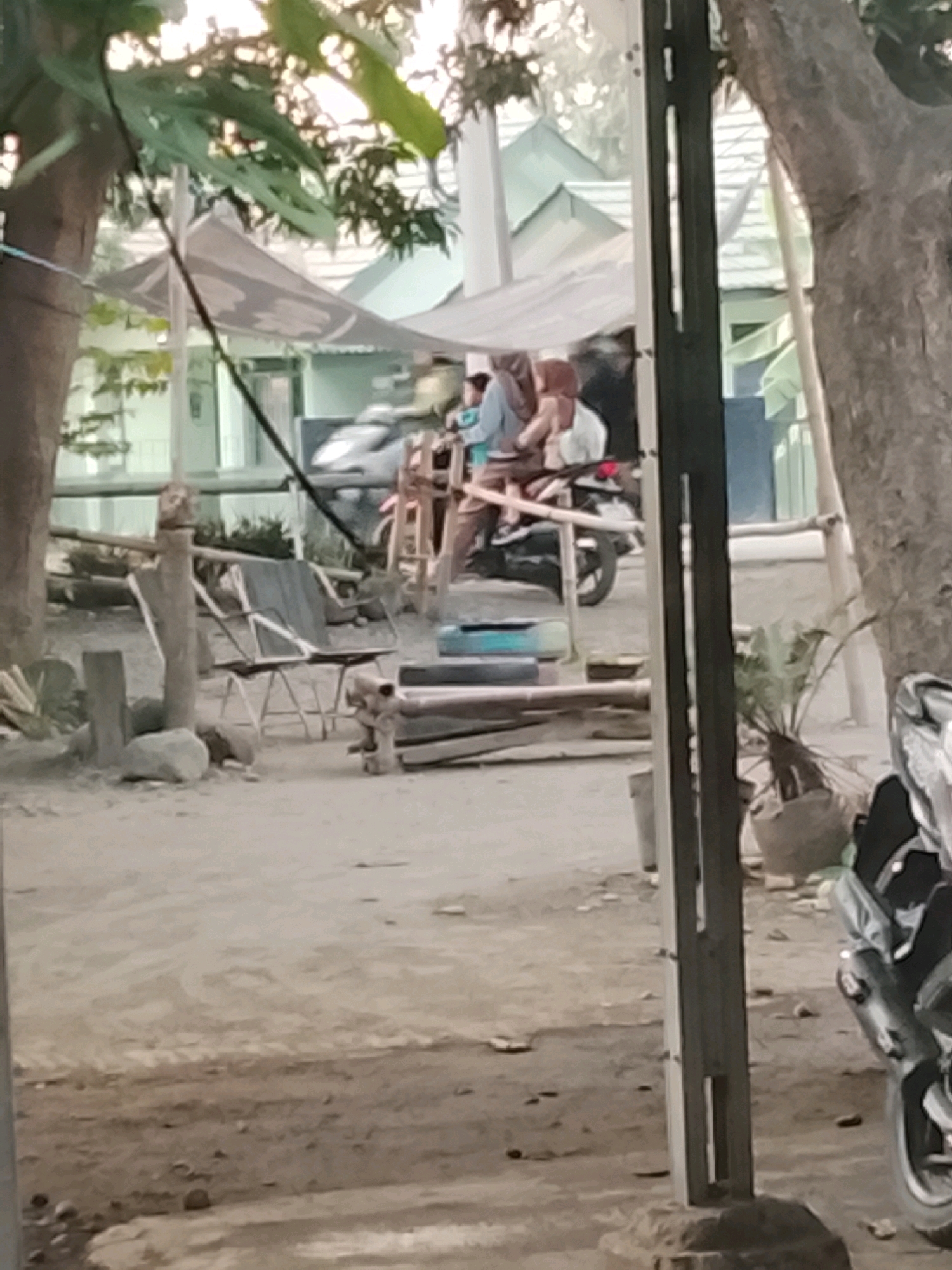 The image depicts an outdoor scene, likely in Ujungrusi, Indonesia, with a dusty, unpaved ground surface. Several elements suggest a residential or communal area. In the foreground, a dark metal pole stands vertically, obscuring part of the view. To the left, a large tree trunk is visible, with leaves and branches overhead partially filtering the sunlight.
In the mid-ground, a motorcycle is parked and partially visible on the right side of the frame. Behind it, several people are seen. Two individuals are seated on the motorcycle, one in the driver's position and another as a passenger, both wearing headscarves and modest clothing. A third person stands behind them. In front of the motorcycle, there is a low wooden structure made of horizontal planks, and to its left, a folded fabric shade is supported by poles.
Further left, a weathered chair with a dark fabric seat and a metal frame is positioned on the ground. Several rocks are scattered around the base of the chair and the area. In the background, several buildings with distinctive sloped roofs are visible, suggesting a settlement. The overall impression is a casual outdoor setting, possibly a residential yard or common area.