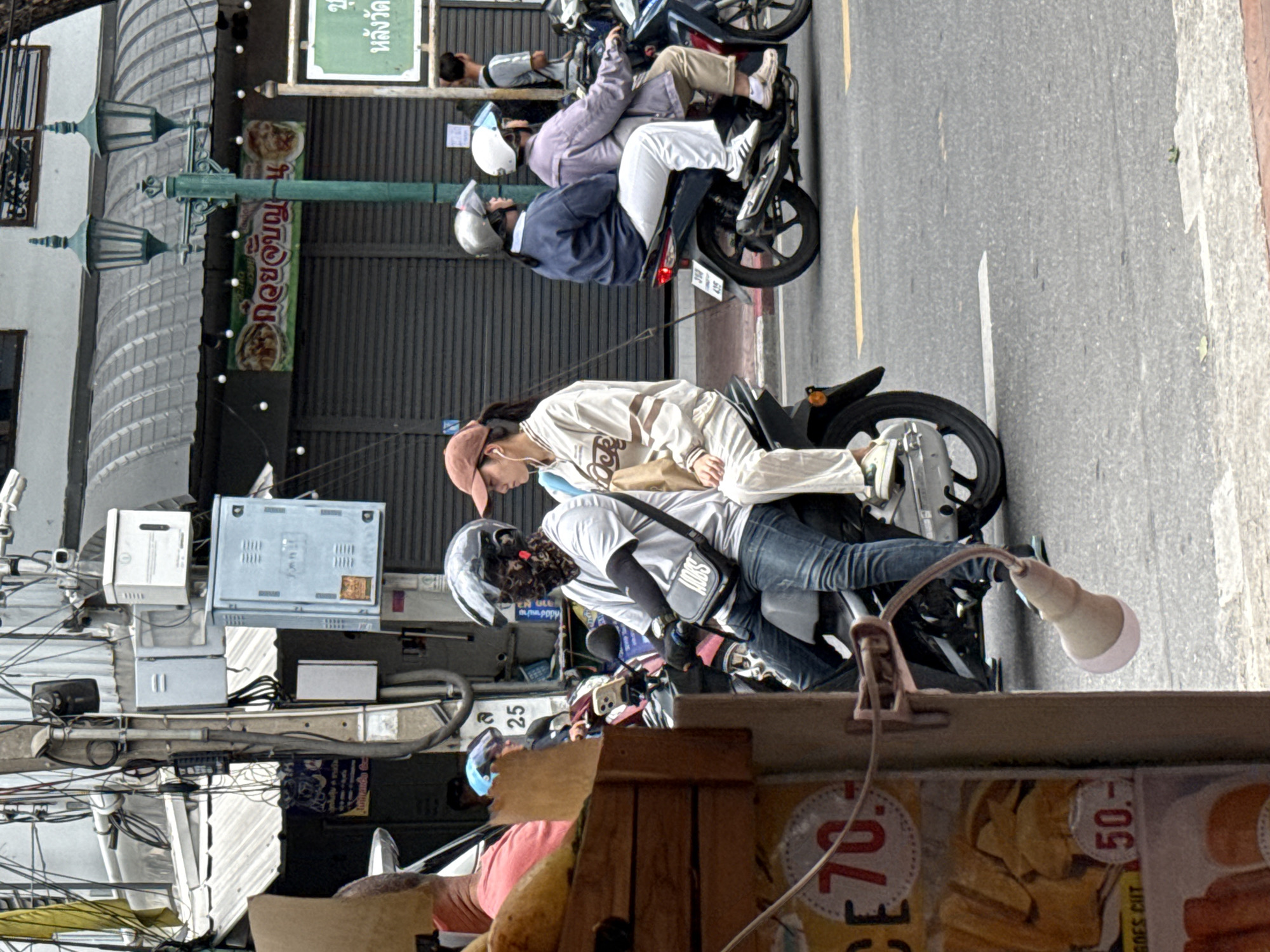 The image shows a street scene in Bangkok, Thailand, with several motorcycles and people. In the foreground, a motorcycle is parked and a person wearing a blue helmet and a pink shirt is partially visible. To the right, two people are riding on a motorcycle. The person in the front is wearing a white helmet and a dark blue shirt, while the person in the back is wearing a light purple jacket and white pants. The rider in the front has a light-colored bag on their lap.
Further to the left, another motorcycle is visible with two people on it. The person in the front is wearing a light-colored shirt and a light brown cap, and appears to be looking to their left. The person behind them is wearing a grey t-shirt and blue jeans, and has a black cloth covering their face. They are carrying a black bag over their shoulder.
In the background, there are buildings with awnings and signs, suggesting a commercial area. One sign has Thai script and images of food. Streetlights and electrical wires are also prominent features of the urban environment. The road surface is asphalt with yellow and white markings. The overall impression is a busy city street with common modes of transportation and commerce.