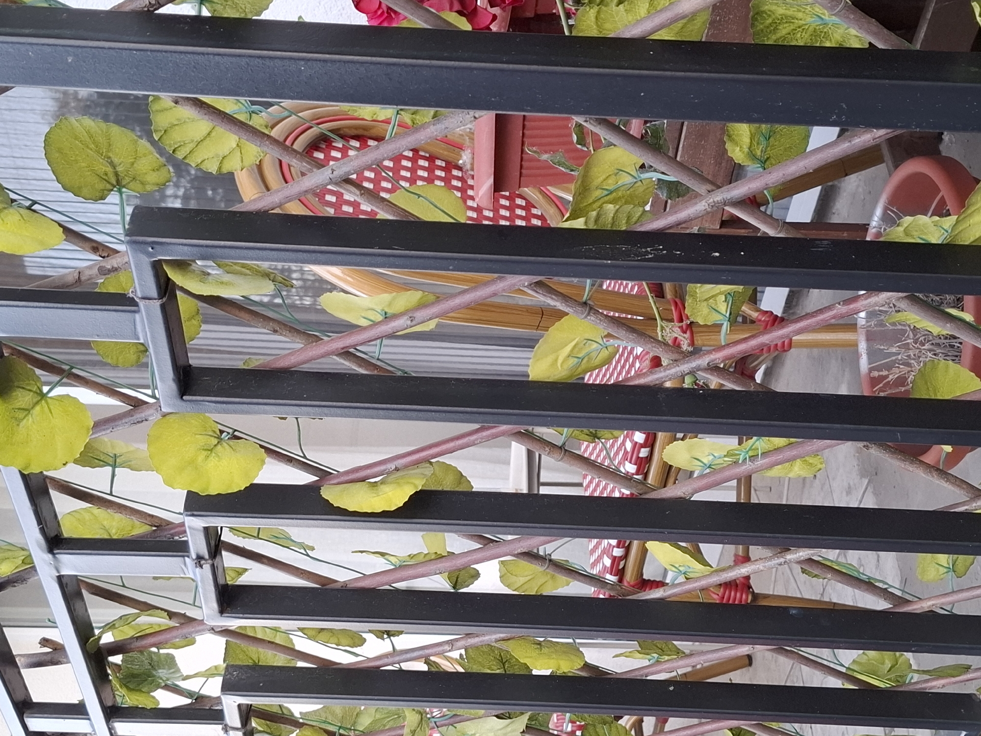 The image depicts a close-up view of a metal structure adorned with artificial foliage, likely part of a pergola or decorative trellis. The structure features black, rectangular metal bars arranged in parallel lines. Woven through the bars are artificial vines with green leaves, adding a touch of nature to the scene. In the background, glimpses of a red and white patterned chair can be observed, suggesting a patio or outdoor setting. The overall impression is of an urban outdoor space with an element of artificial greenery. There is no explicit evidence in the image to pinpoint the exact location within Zemun Urban Municipality, Serbia.