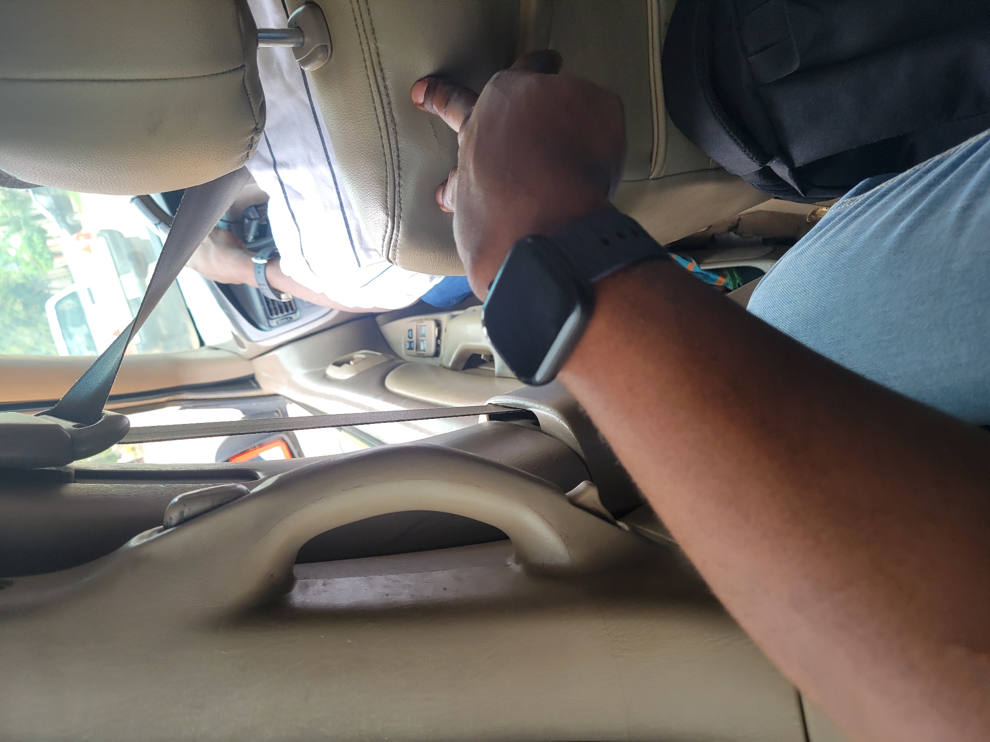 A first-person perspective shot captures a person inside a vehicle, likely a car, in Katambe, Nigeria. The view is directed towards the front of the vehicle, with the driver's seat visible. A person in the driver's seat, wearing a white shirt with thin black stripes, is holding the steering wheel and appears to be driving. A seatbelt is fastened across their chest. The interior of the car is a light tan color, featuring a textured upholstery on the seats and door panels. A black smartwatch is visible on the left wrist of the person taking the photo. In the background, through the car window, a street scene is discernible, with a white car and trees suggesting an urban or suburban environment. The lighting indicates it is daytime.