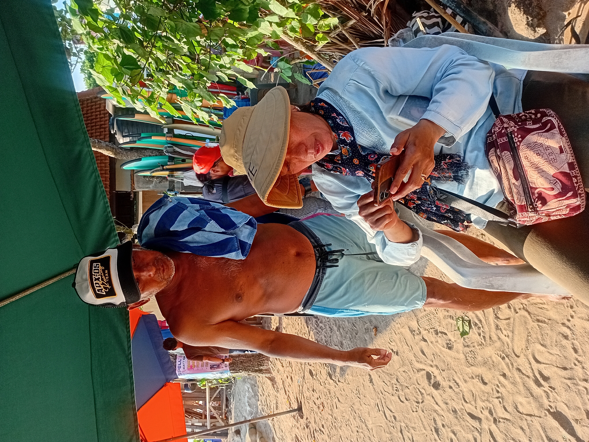 In Kuta, Indonesia, on a sandy beach, two individuals are captured in a moment. A shirtless man wearing a black and white trucker hat and light blue swim trunks stands to the left, a striped towel draped over his shoulder. He is looking down, appearing to be in conversation or observing something. To the right, a woman with a wide-brimmed hat and a blue patterned scarf sits, engrossed in her smartphone. She is wearing a light blue long-sleeved shirt and has a patterned shoulder bag with elephant motifs. Behind them, a collection of surfboards are propped up, suggesting a surf school or rental shop. Umbrellas and beach structures are visible in the background, indicating a bustling beachside location. The lighting suggests it is late afternoon or early morning, with the sun casting warm tones on the sand and people.