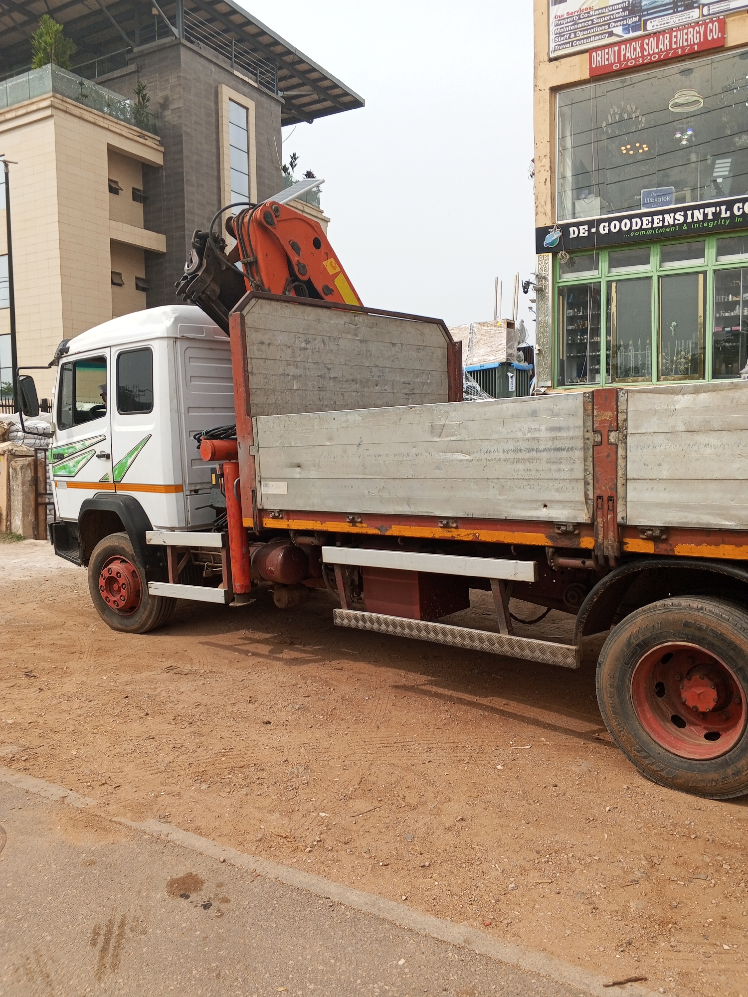 The image depicts a white medium-duty flatbed truck with an orange crane mounted behind the cab, parked on a reddish-brown dirt surface near a paved road. The truck's wheels are red, and the flatbed has a weathered silver color with orange accents. In the background, there is a multi-story building with a light-colored facade and a modern design, including visible vegetation on a rooftop terrace. To the right of the truck, a commercial building with a large glass storefront displays signs for "ORIENT PACK SOLAR ENERGY CO." and "DE-GOODEENS INT'L CO." The scene appears to be in a commercial or industrial area. The presence of the named businesses and the location of the city of Abuja, Nigeria are mentioned.