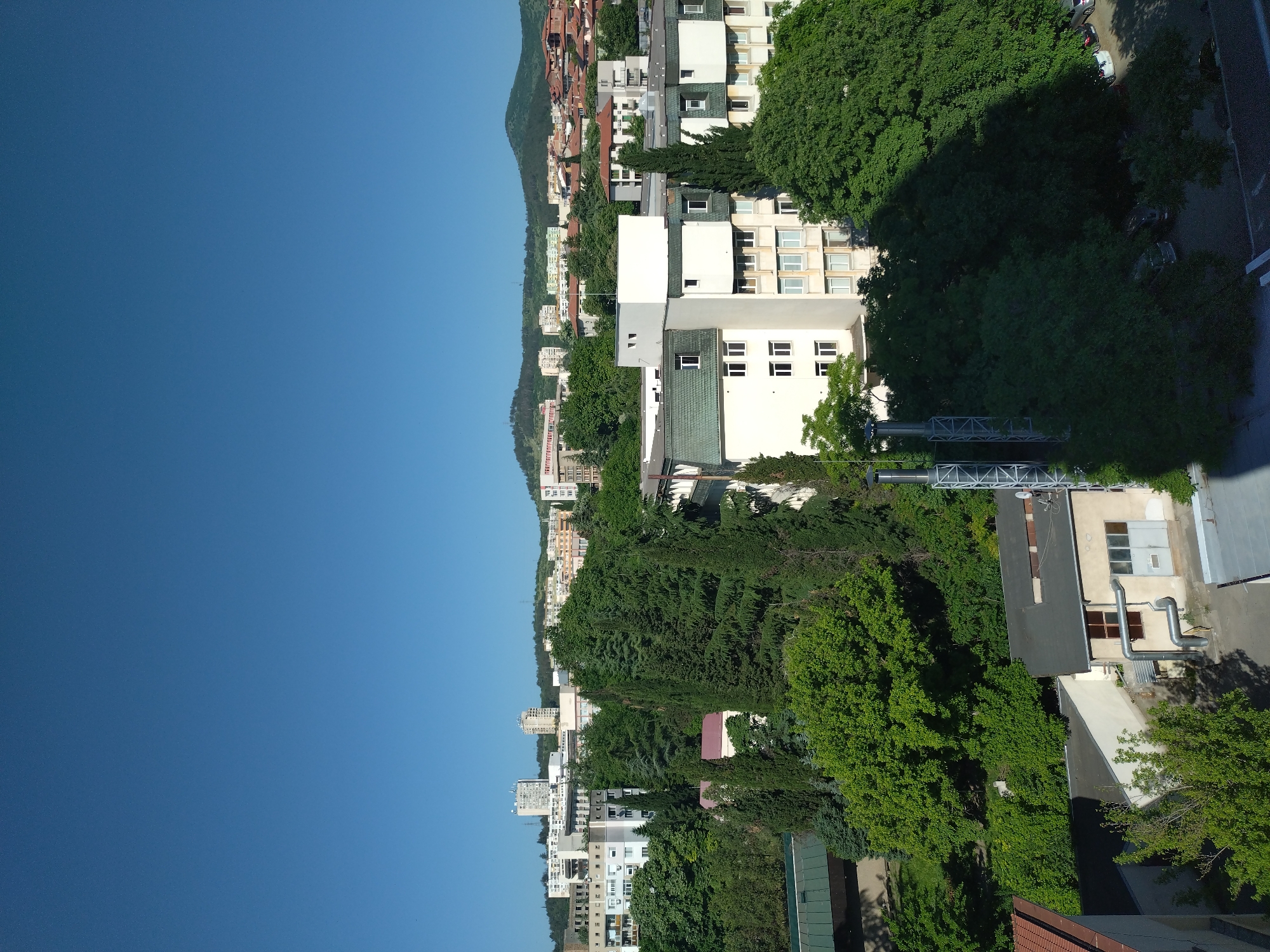 This is an aerial view of a neighborhood in Stara Zagora, Bulgaria on a sunny day. The sky is clear blue. The scene is dominated by buildings of various heights and styles, mixed with dense green trees. Along the left side of the image, a line of buildings runs along the base of a hill or low mountain, with vegetation covering the hillside. The buildings closer to the foreground appear to be apartment complexes, while buildings further into the distance are smaller and more scattered. In the foreground, on the right side of the image, is a commercial or industrial building with a prominent metal chimney-like structure. A road or street runs along the edge of the buildings on the right side of the frame, partially obscured by tree cover. No people or animals are visible in the image.