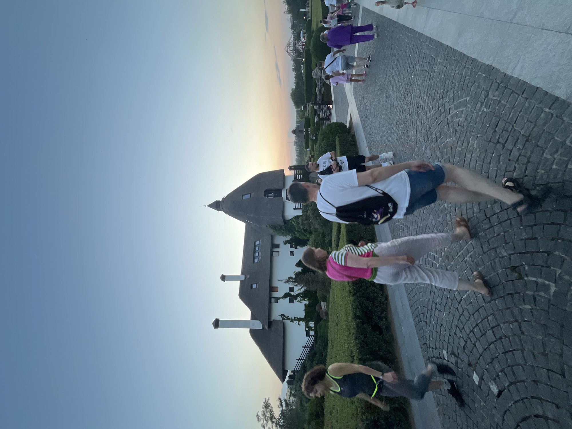 The image shows an outdoor scene at dusk, likely in a park or recreational area, in the city of Dnipro, Ukraine. Several people are walking on a cobblestone path in the foreground. To the left, a woman with curly dark hair and a neon green trimmed athletic top walks past a meticulously manicured hedge. Next to her, a woman with light brown hair wears a bright pink vest over a striped shirt and light-colored pants. In the center foreground, a man with a backpack walks away from the camera, wearing a white t-shirt and denim shorts. Further back, other individuals and a goose are visible on the path and surrounding area. The background features a distinctive building with a thatched roof and white walls, reminiscent of European architectural styles, along with trees and other structures, including what appears to be a Ferris wheel or similar amusement park ride in the distance. The sky is a pale blue with hints of orange and pink from the setting sun. The overall impression is of a tranquil evening outing in a well-maintained public space.