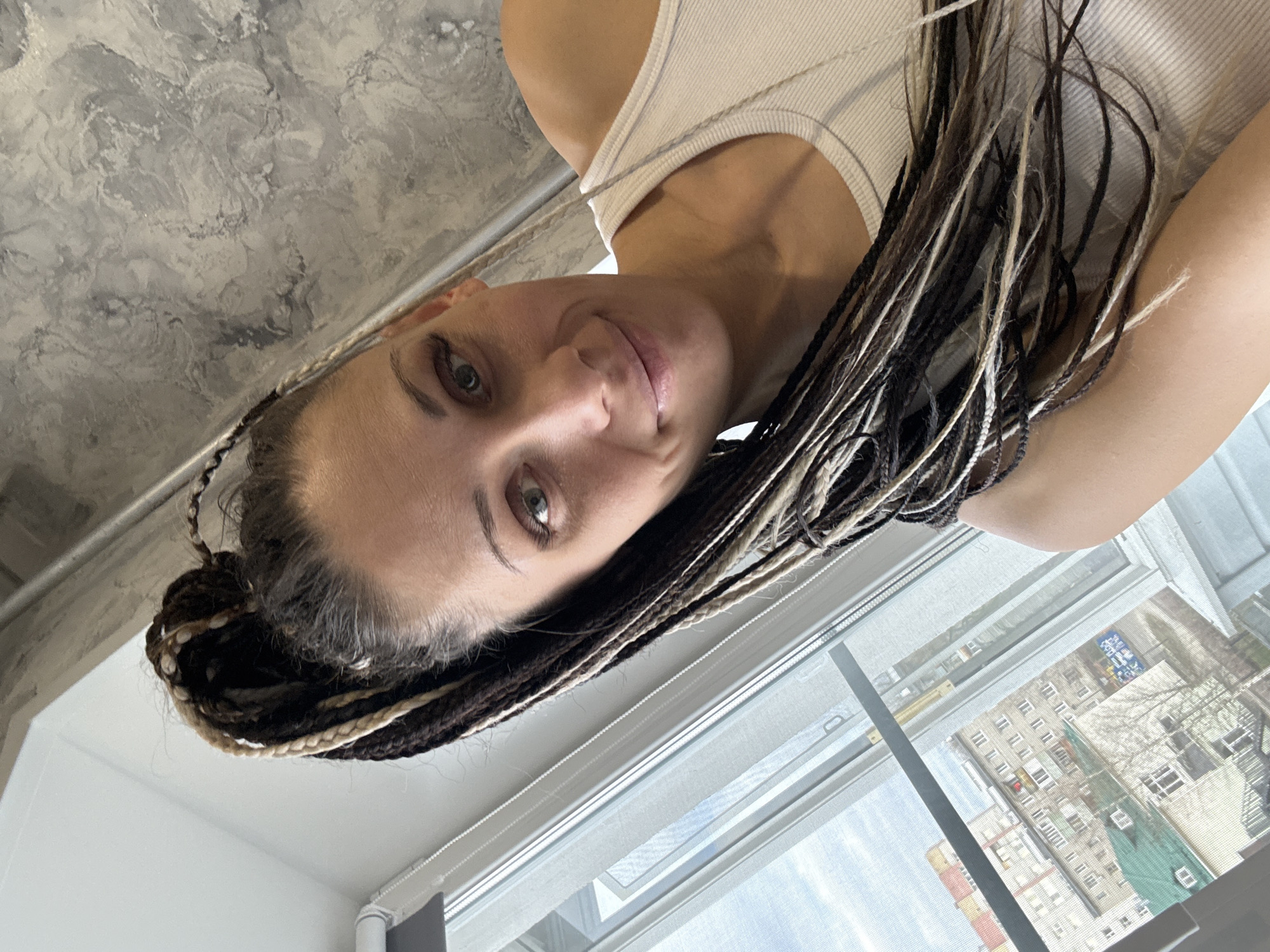 A woman with multi-colored dreadlocks, featuring dark brown and blonde strands, is captured in a selfie taken from an upward angle. Her head is tilted, and she looks directly at the camera with a soft, subtle smile. She has light-colored eyes, possibly green or hazel, and is wearing a light-colored, ribbed tank top.
The setting appears to be an indoor space. Above her head, a textured, mottled gray and white ceiling, resembling concrete or decorative plaster, is visible. To her right, a large window reveals an urban landscape. Outside the window, several apartment buildings are visible, indicative of a city environment. One building features a distinctive green roof or tarp, and another has a blurry blue sign with some indistinct text. Bare trees suggest the season is likely winter or early spring. Blinds are partially drawn on the window, and the bright light streaming in confirms it is daytime. The scene is located in Rusanovo, Russia. The woman's expression suggests a calm and relaxed demeanor.