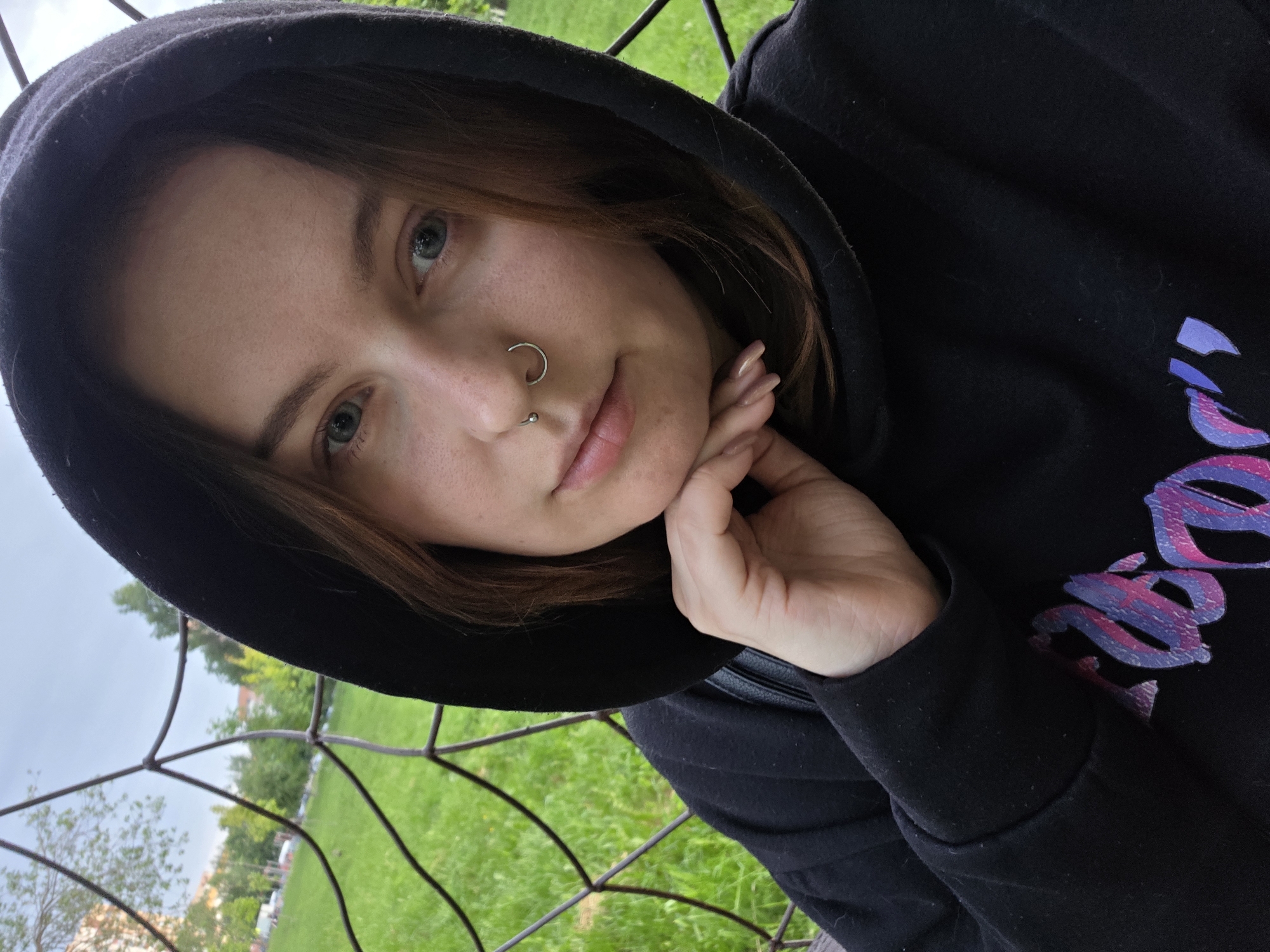 A young adult female, estimated to be in her late teens or early twenties, is depicted in a chest-up portrait. Her light-colored eyes, possibly blue or grey, are directed at the viewer. Brown hair is visible beneath a raised black hoodie. Her complexion is fair, with visible freckles. She has two silver facial piercings: a septum ring and a nostril stud on the viewer's right nostril. Her lips are naturally pigmented. She wears a black hooded sweatshirt with a partially visible graphic design in shades of purple and pink on the chest. Her right hand, with light-colored fingernails, supports her chin. The background consists of a wire fence or netting in the foreground, beyond which lies a grassy park or field with green trees. Distant urban architecture is indistinctly visible. The sky is overcast. The location is an outdoor park environment within the city of Unknown, Unknown.