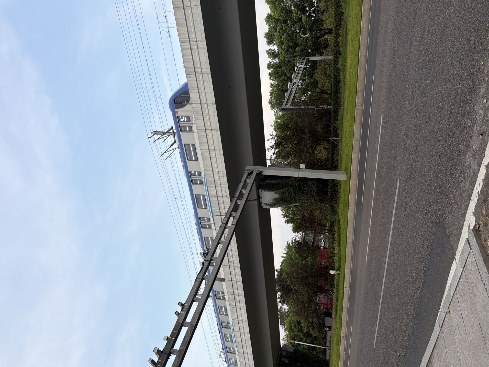 An elevated train track spans across a highway with a train in motion. The train is light gray with blue accents. The track is supported by concrete pillars. A patch of green grass and trees line the side of the highway. The sky is light blue with some clouds. The image appears to be taken from the perspective of someone on the side of the highway, looking up at the train.