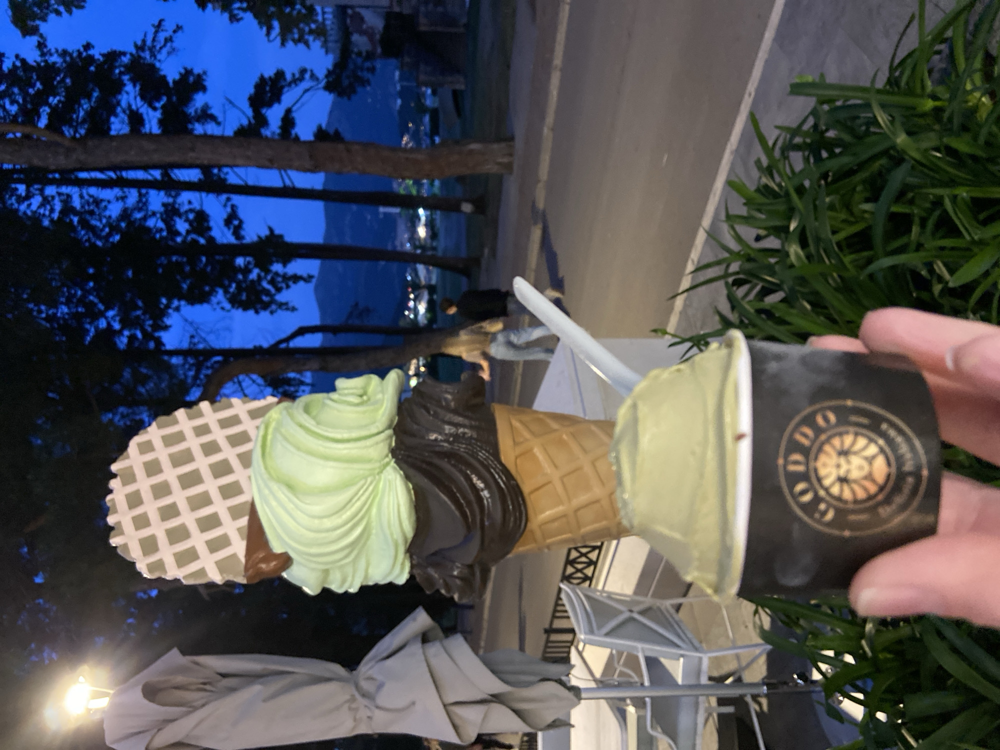 The image shows a hand holding a small cup of pistachio-colored gelato with the logo "GODDO" on it. Next to the cup is a large, decorative, multi-scooped ice cream cone placed next to outdoor seating under umbrellas. In the background, there are trees, a street with people walking, and mountains with city lights. Based on the logo on the gelato cup, the photo was taken in Budva, Montenegro.