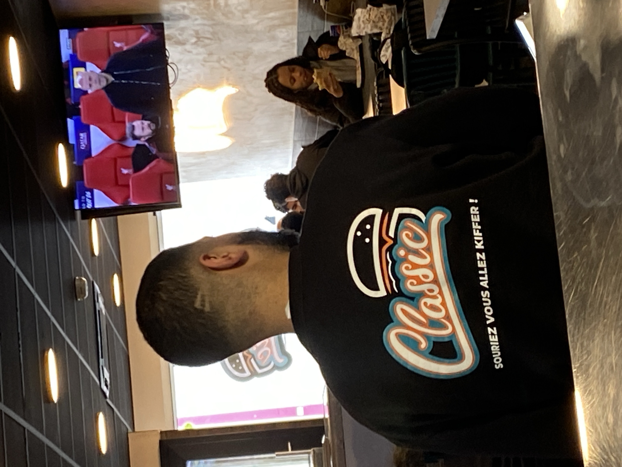 The image shows the interior of a restaurant in Schiltigheim, France. A man wearing a black sweatshirt with a "Classic" burger logo is seated at a table. A woman with long hair is eating a sandwich at another table. A television is mounted on the wall, displaying a soccer match. The interior features a dark wall with vertical lights and a window.