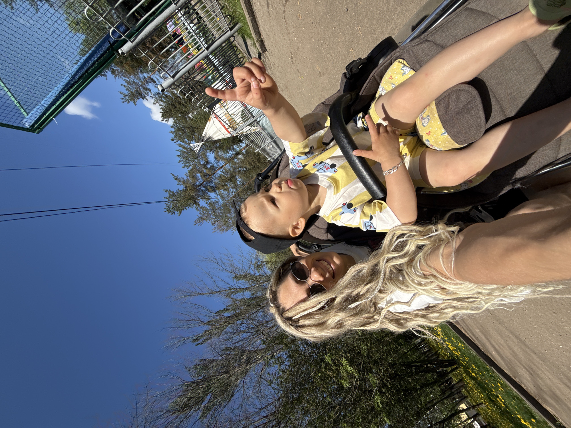 A sunny outdoor shot shows a woman with long, blonde, braided hair wearing sunglasses, and a toddler in a stroller. The child is wearing a yellow and white patterned shirt and has a black cap on. They are holding onto the stroller's bar with one hand and raising the other hand. The woman is smiling and looking at the camera. The background includes trees, a blue sky with a few clouds, a fence, and some yellow flowers. The location is a city park.