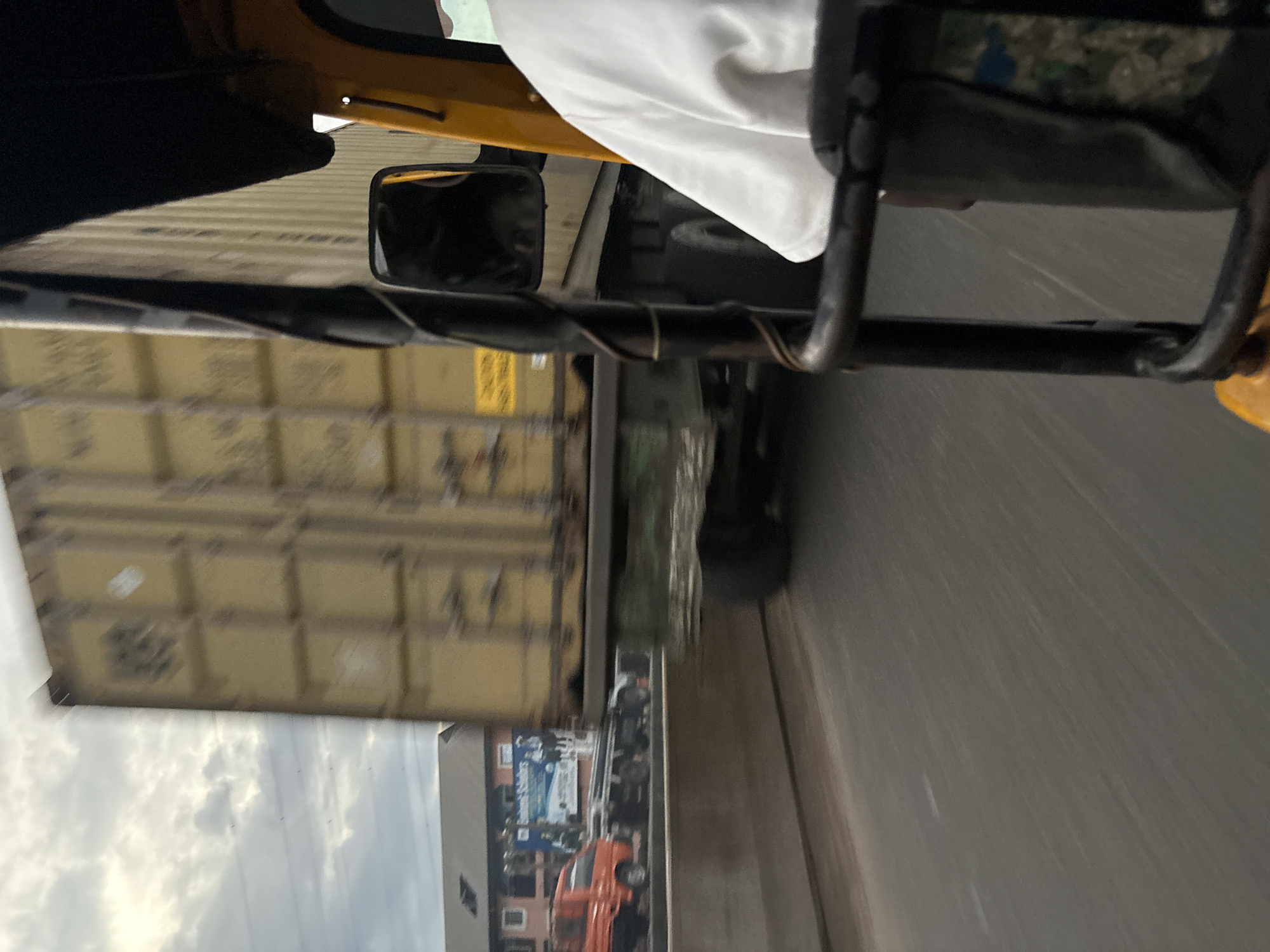 The image depicts a street scene in Onitsha, Nigeria, captured from the perspective of a moving auto-rickshaw. In the foreground, the side of the auto-rickshaw is visible, including a black metal frame and a portion of a yellow body. A person wearing a white shirt is partially visible inside the auto-rickshaw, suggesting they are a passenger.
Dominating the mid-ground is a large, yellow shipping container being transported on a truck. The truck and its cargo are significantly blurred due to motion, indicating the vehicles are traveling at speed. The container has visible grid-like markings and some faint logos or text.
In the background, several elements of the urban environment are discernible. To the left, a multi-story building with a reddish facade and multiple windows can be seen. In front of this building, a smaller, orange truck is parked or moving slowly. A billboard with text that includes "Business Scholars" and images of people is also visible behind the orange truck. The sky above is cloudy, with a mixture of light and dark grey clouds. The road surface is asphalt and shows motion blur. A concrete barrier separates the road from what appears to be a lower level or embankment. The overall impression is one of bustling city traffic.