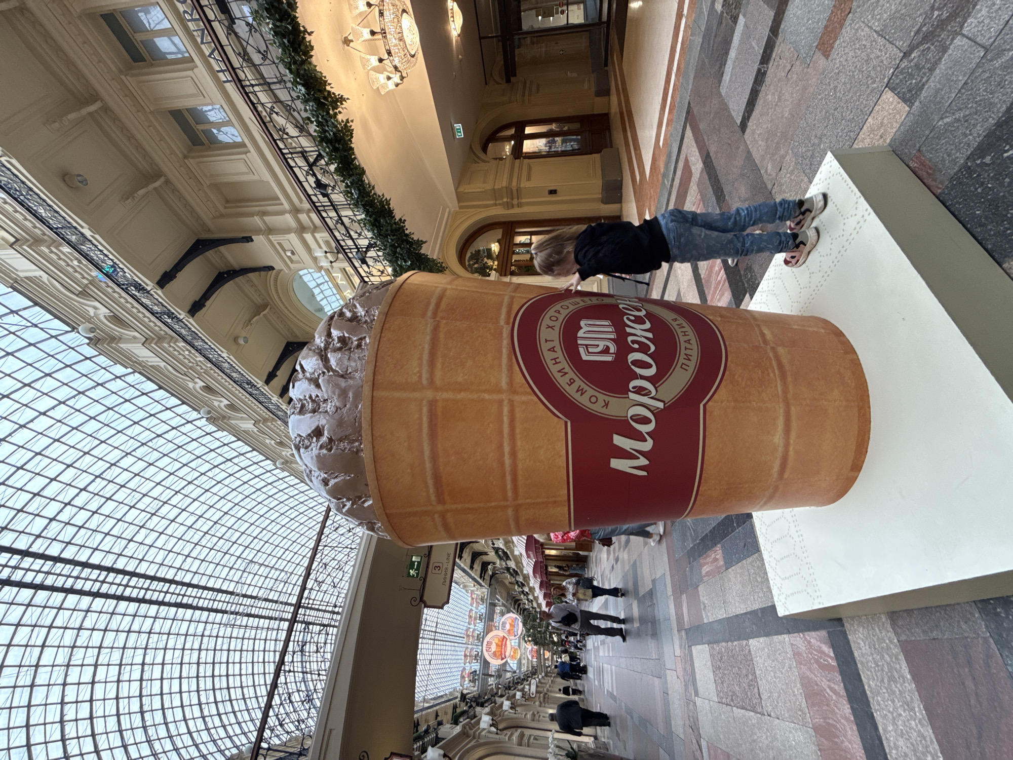 The image depicts a large, oversized ice cream cone statue, likely a prop for a retail establishment. The statue is positioned in the foreground, to the right of center. A young girl, wearing a black hoodie and blue jeans, is standing next to the statue, with her hands touching its surface. She is facing away from the viewer, looking up at the ice cream. The statue itself is a representation of a chocolate ice cream cone with a textured waffle cone. A circular logo with Russian text is visible on the cone, reading "ГУМ" and "МОРОЖЕНОЕ" (ice cream).
The background reveals an interior of a grand, spacious building with a distinctive arched glass ceiling, characteristic of the GUM department store on Red Square in Moscow, Russia. The architecture features multiple levels with balconies, ornate railings, and chandeliers. People can be seen walking in the distance along a wide, tiled walkway lined with shops. The overall environment suggests a busy, upscale shopping mall or department store. The flooring is a mosaic of rectangular and square tiles in various shades of grey, pink, and black. The lighting appears to be a mix of natural light filtering through the glass ceiling and artificial light from chandeliers and ceiling fixtures.