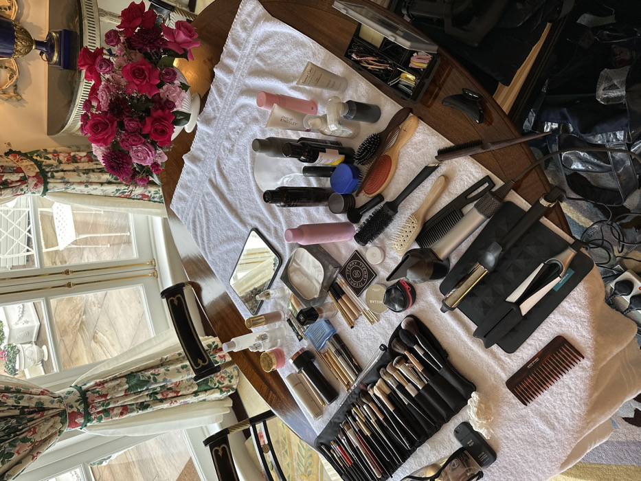 The image captures an indoor scene, likely within a residence or hotel in London, United Kingdom. A large, polished dark wooden table occupies the foreground and midground, partially covered by a white terry cloth towel. The table is extensively arranged with beauty and grooming products, indicating a professional or elaborate styling setup.
On the towel, a black roll-up case displays a comprehensive collection of makeup brushes. Adjacent to this are numerous cosmetic items, including bottles, jars, tubes, compacts (with visible branding such as By Terry and Chanel), a small rectangular handheld mirror, and an eye mask. A variety of hair styling tools are also present: multiple hairbrushes (paddle, round, vent), various combs, a hair straightener, a curling iron, and a hairdryer nozzle. Hair clips and pins are systematically organized in a multi-compartment clear tray.
A large floral arrangement, composed of dense red and pink roses, dahlias, and other blossoms, is displayed in a white classical urn-style vase on the table.
The background features tall, multi-paned windows with gold-colored sashes, offering a view of an outdoor paved patio area furnished with white potted plants and white metal chairs. The windows are framed by ornate floral-patterned curtains (red, pink, and green on a cream background), tied back with green sashes. To the right of the floral arrangement, an ornate gold-framed piece (likely a mirror or artwork) hangs above a small decorative side table holding a blue and gold urn. Electrical cords from the hair tools lead to a power strip visible on the floor beneath the table, alongside dark bags. No individuals are present in the scene.