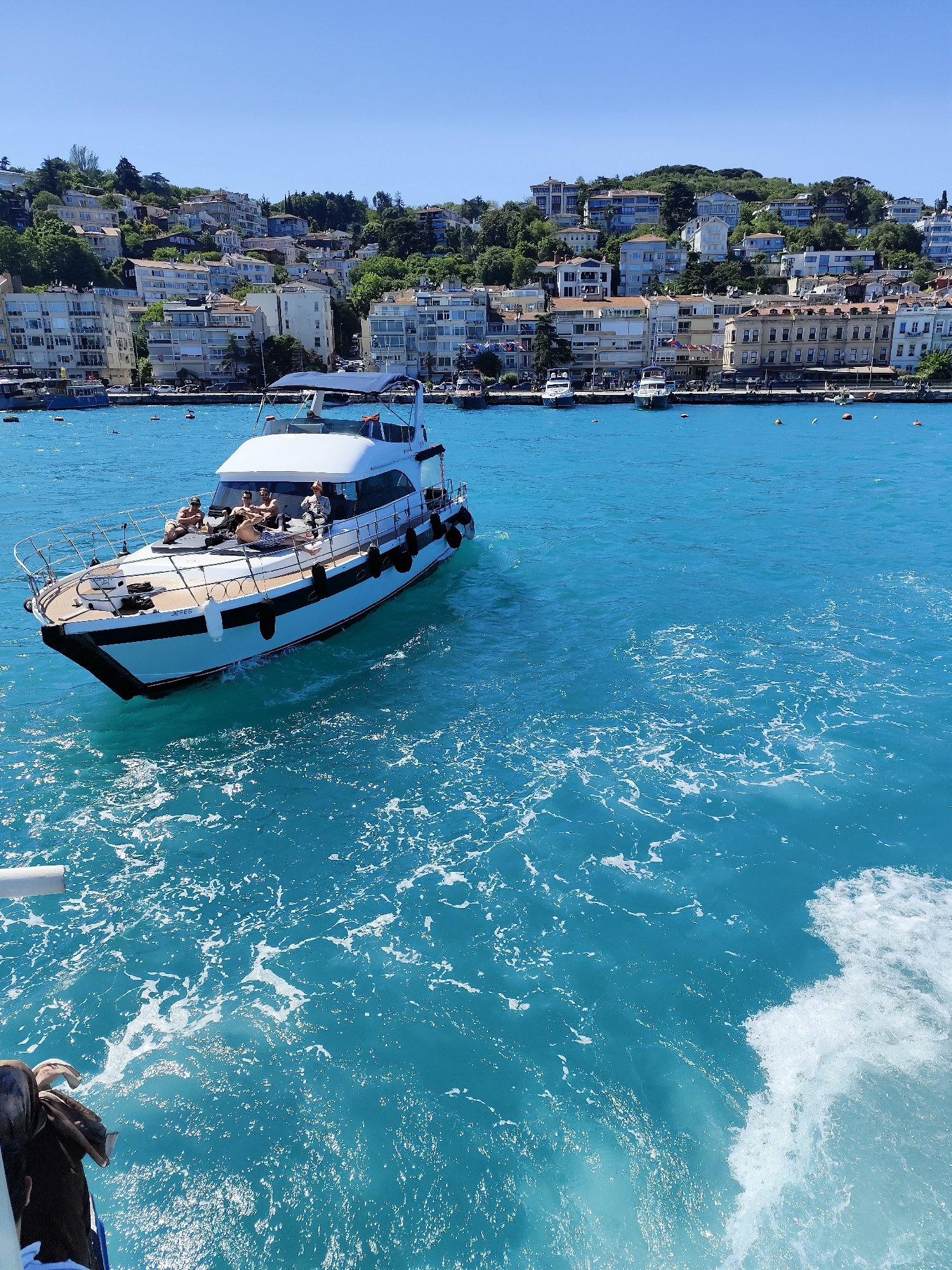 Captured during daylight on a bright, sunny day in Beşiktaş, Turkey, the scene shows a white yacht occupied by at least four people navigating the bright turquoise waters of the Bosphorus strait. The yacht features a black and white hull, a wood deck, and black fenders along its sides. The people are casually positioned on the front deck.
In the background, a densely populated hillside is visible, displaying multi-story residential and commercial buildings arranged along the shoreline. The buildings are diverse in style, with tan and white colors predominating. Trees are interspersed among the buildings, contributing to the urban landscape.
The water shows signs of movement and wake patterns indicating the yacht is underway. The viewpoint is slightly elevated and tilted, presumably taken from another vessel or a high vantage point. The color and clarity of the water suggest a sunny day and clear visibility. Small boats and buoys are scattered across the sea in the background, suggesting maritime activity and the location's function as a harbor or waterway.