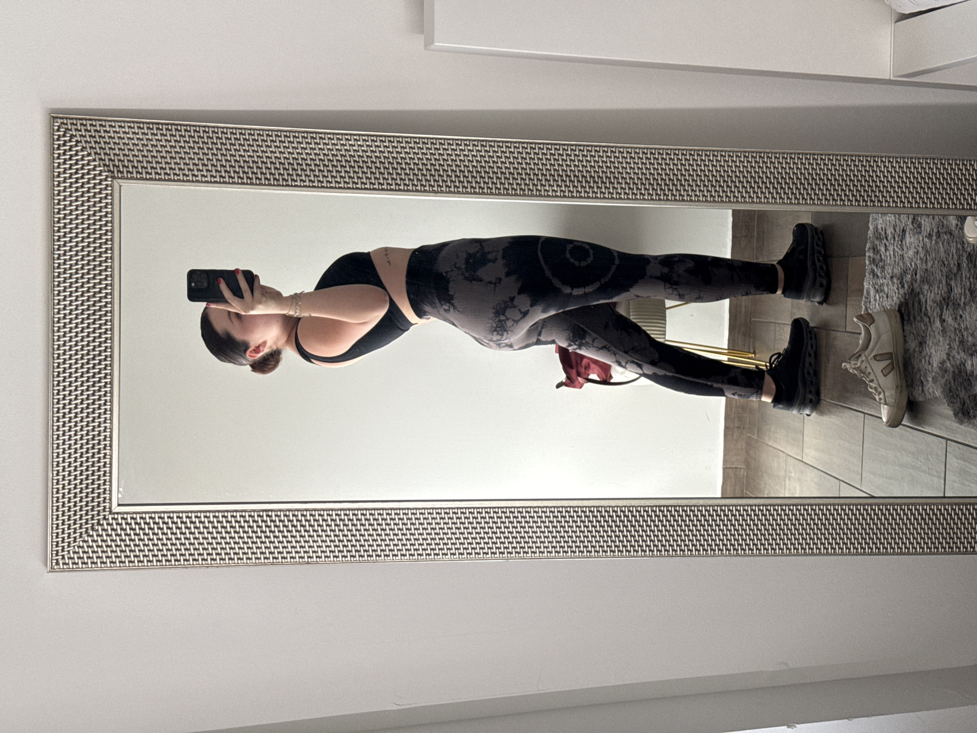 A person is seen in a full-length mirror, taking a selfie. The individual, from the chest down to the feet, is wearing a black sports bra and dark grey leggings with a black tie-dye pattern. They are holding a black smartphone in their right hand to capture the image, with their jawline, ear, and hair pulled back into a bun visible. Red nail polish is noticeable on their fingers.
The person is wearing black athletic shoes. On the light-colored tiled floor to their right, a grey textured rug is visible, upon which another pair of light-colored sneakers rests next to the person's feet. The mirror itself has a distinctive, wide frame featuring a woven metallic pattern in shades of brown and silver. The background reflected in the mirror consists of plain white or light grey walls, suggesting an indoor setting with ample ambient light, likely during the daytime. No discernible text is visible in the image.