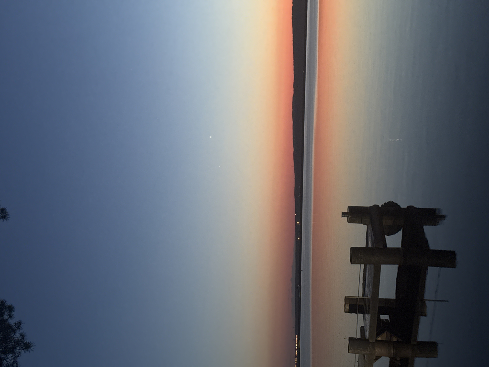 This image depicts a serene twilight or sunset scene over a calm body of water in Chaikovsky, Russia. The sky transitions from a deep, dark blue at the top, where dark, silhouetted pine branches are visible in the upper left corner, to a lighter blue mid-sky. Two distinct bright points, likely planets or bright stars, are visible in the lighter blue portion of the sky. Near the horizon, the sky displays a vibrant band of orange and red, reflecting the setting sun. A dark, undulating landmass, possibly a distant shore or hills, separates the sky from the water. On the far left of this landmass, faint artificial lights are discernible. The water body, appearing smooth and tranquil, perfectly mirrors the sky's gradient colors and the dark landmass. In the bottom foreground, a dark, low-lying pier or dock structure, constructed of several concrete or wooden posts supporting a platform, is partially submerged. This structure casts a clear, dark reflection onto the water. No individuals or active subjects are present in the scene.
