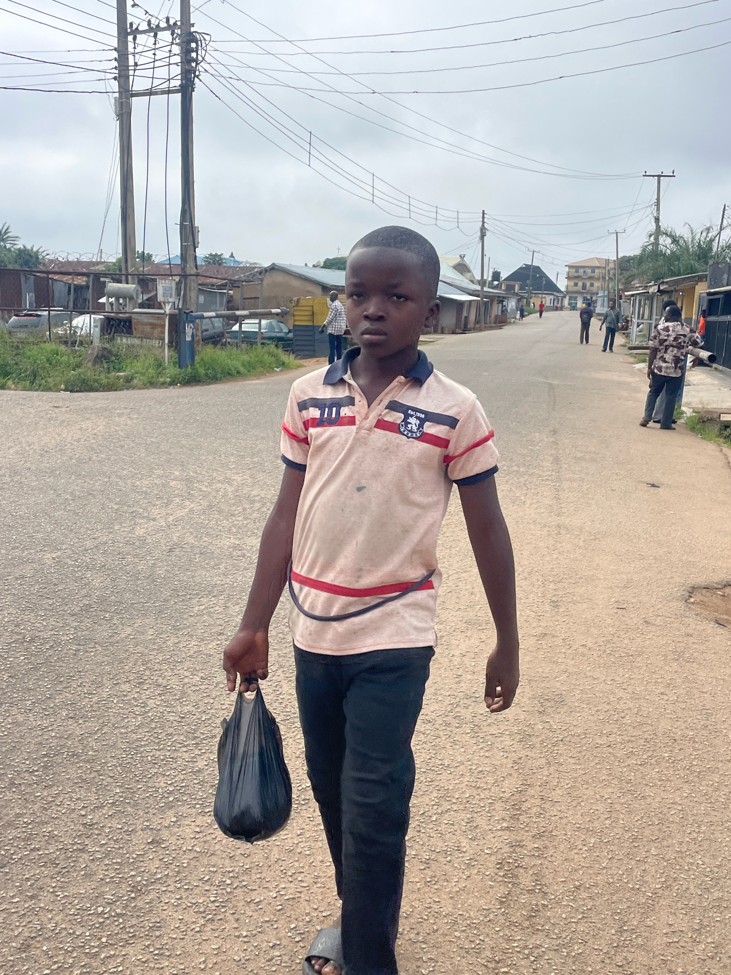 A young boy walks down a paved street in Jos, Nigeria, carrying a black plastic bag in his left hand. He is wearing a light pink polo shirt with navy blue and red accents, and dark pants. His gaze is directed forward, and his expression is neutral. The street is lined with buildings and utility poles with numerous wires crisscrossing overhead. Several other people are visible in the background, some walking along the street and others standing. The sky is overcast, suggesting a cloudy day. The pavement appears to be a mix of asphalt and gravel.