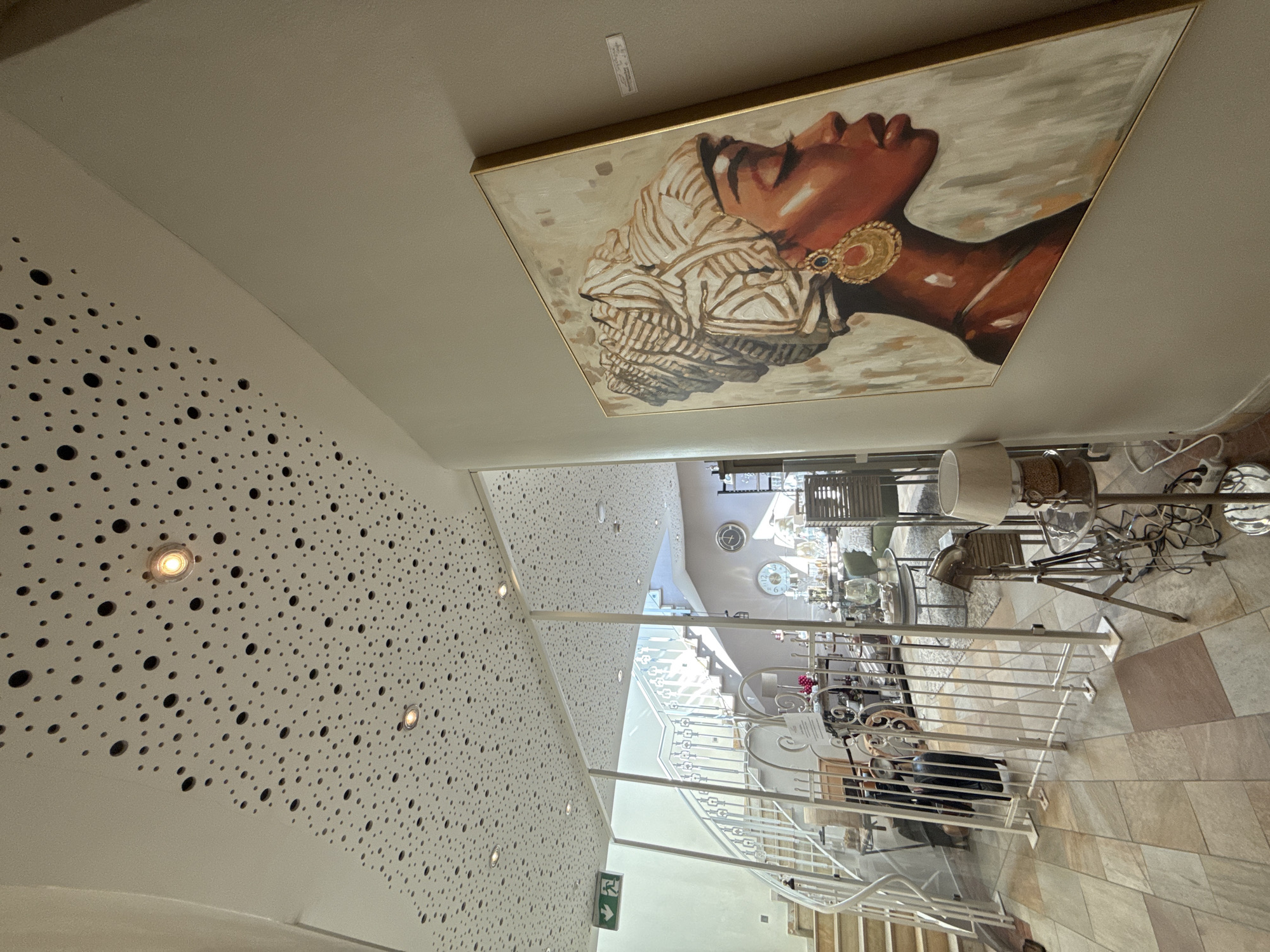 This image shows the interior of a shop in Bad Sachsa, Germany. The shop features a white perforated ceiling with numerous circular openings, some of which are illuminated by spotlights. A large painting of a woman wearing a headwrap and large earrings hangs on a light beige wall to the right of the frame. In the background, a white staircase with decorative railings is visible, along with various retail displays containing decorative items and furniture. A green emergency exit sign is mounted on the left side of the frame. The floor is tiled with large, irregularly shaped tiles in earthy tones. A lamp on a metallic stand is positioned in the foreground. The overall impression is that of a well-lit, modern retail space showcasing decorative art and home furnishings.