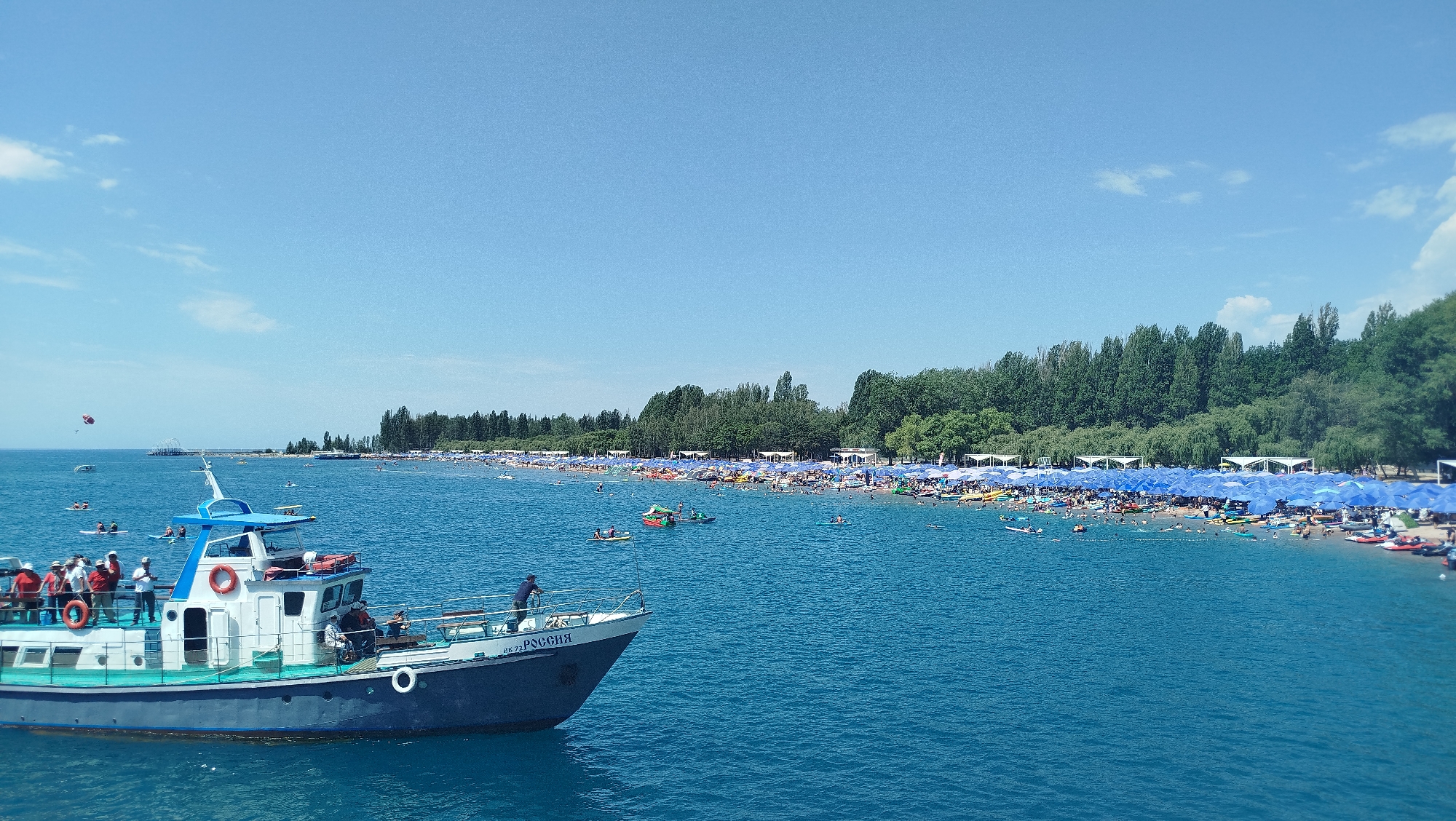 A wide shot captures a bustling beach scene under a clear blue sky. In the foreground, a white and blue boat with several people on deck sails on bright blue water. The boat has a visible nameplate on its hull that reads "UK-72 РОСSIЯ". In the midground, a crowded beach stretches along the shoreline, dotted with numerous blue umbrellas providing shade. People are seen swimming, sunbathing, and engaging in various water activities. The background features a dense line of green trees, separating the beach from the distant landscape.