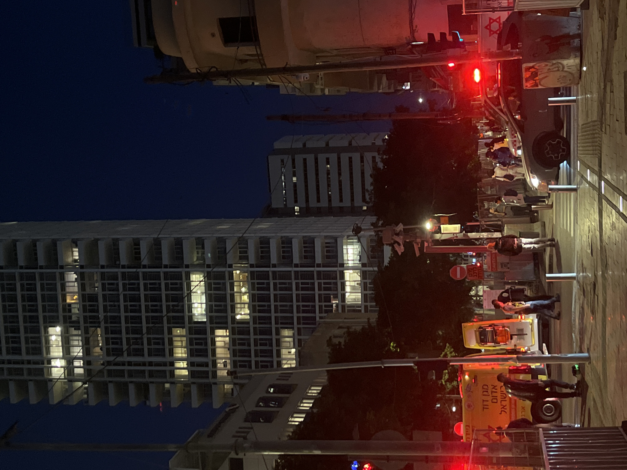The image captures a nighttime scene in Tel-Aviv, Israel. A tall, modern building with many illuminated windows dominates the left side of the frame. To the right, there's a street scene with an ambulance parked. People are standing and walking around the ambulance. Streetlights and traffic signals are visible, adding to the urban setting. A vehicle with what appears to be a Star of David on it is parked on the sidewalk nearby.