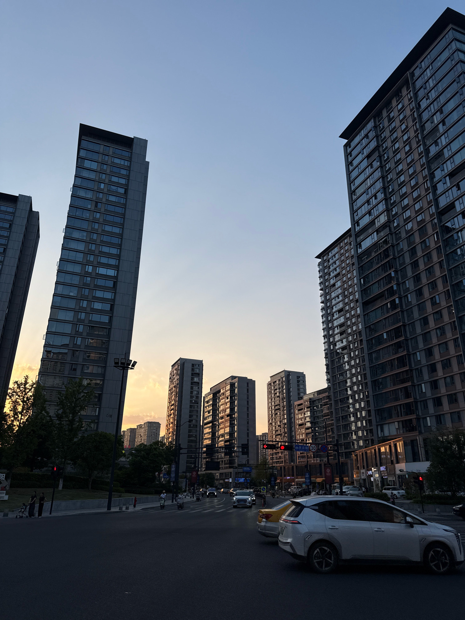 The image depicts a street scene in Yiwu, China, at dusk. Tall residential buildings with numerous windows dominate the skyline. The setting sun casts a warm glow, visible through the gaps between the buildings and reflected on their facades. A street intersection is in the foreground with cars and scooters navigating through traffic. Pedestrians are visible on the sidewalks, and traffic signals are illuminated. The sky is clear with a gradient of orange and blue from the sunset.
