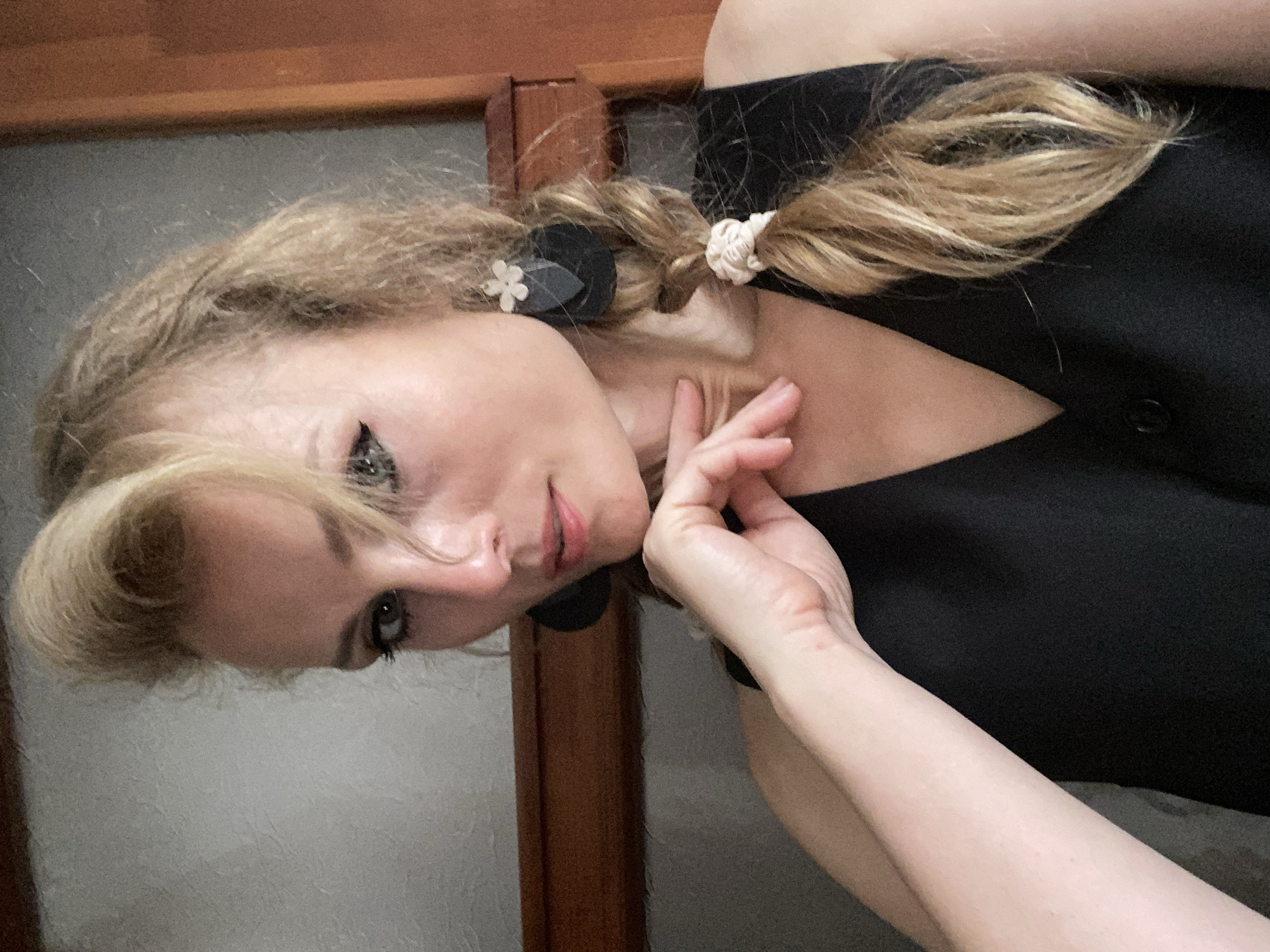 The image shows a woman with blonde hair pulled into a braid. She is wearing a black vest and large black earrings with a white flower accent. Her left hand is resting on her chin. The background appears to be an indoor setting with a wooden shelf and a textured wall. There is no indication of a specific location within the city of Lida, Belarus.