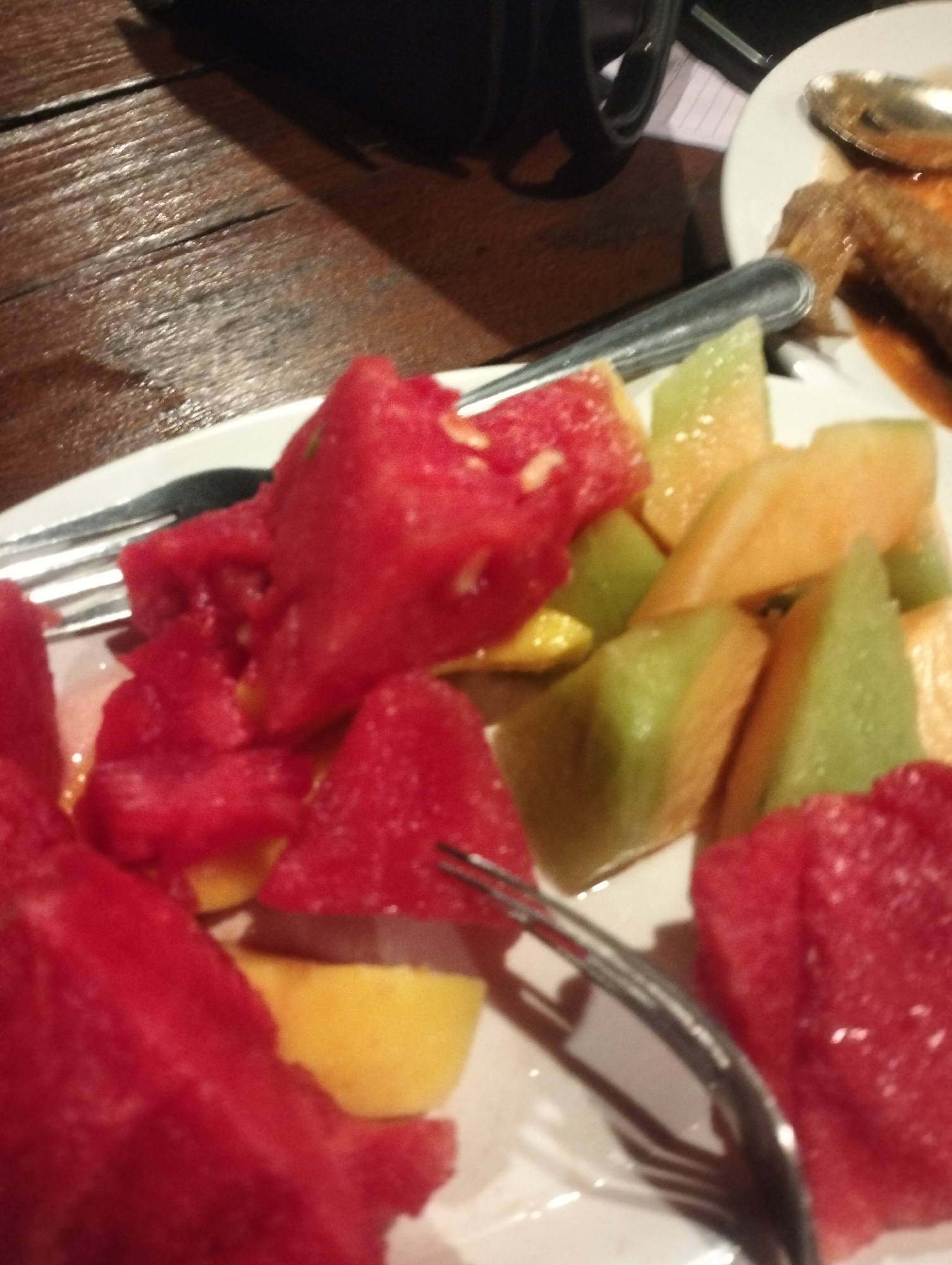 A close-up, slightly blurry image captures a plate of freshly cut fruit and part of another dish on a dark wooden table. The main subject is a white ceramic plate filled with an assortment of fruit cubes, predominantly bright red watermelon. Interspersed with the watermelon are chunks of orange cantaloupe and pale green honeydew melon. A silver-colored metal fork is resting on the plate, partially covered by the fruit, with its tines pointing towards the bottom right. Another fork or knife is visible on the top left edge of this plate.
In the upper right corner, a portion of a second white plate is visible, holding a savory dish, which appears to be pieces of meat (possibly chicken or fish) covered in a rich, dark brown sauce. A silver spoon is resting on this plate. The table itself is made of dark brown wood with a visible grain pattern, indicating an indoor setting. In the blurry background, a black object, potentially a camera or a cup, and a white piece of paper with faint lines are discernible. The scene depicts a meal in progress or prepared for consumption, likely in Keudekruenggeukueh, Indonesia. No specific activity beyond dining is shown, and no text is visible. The lighting suggests an indoor environment.