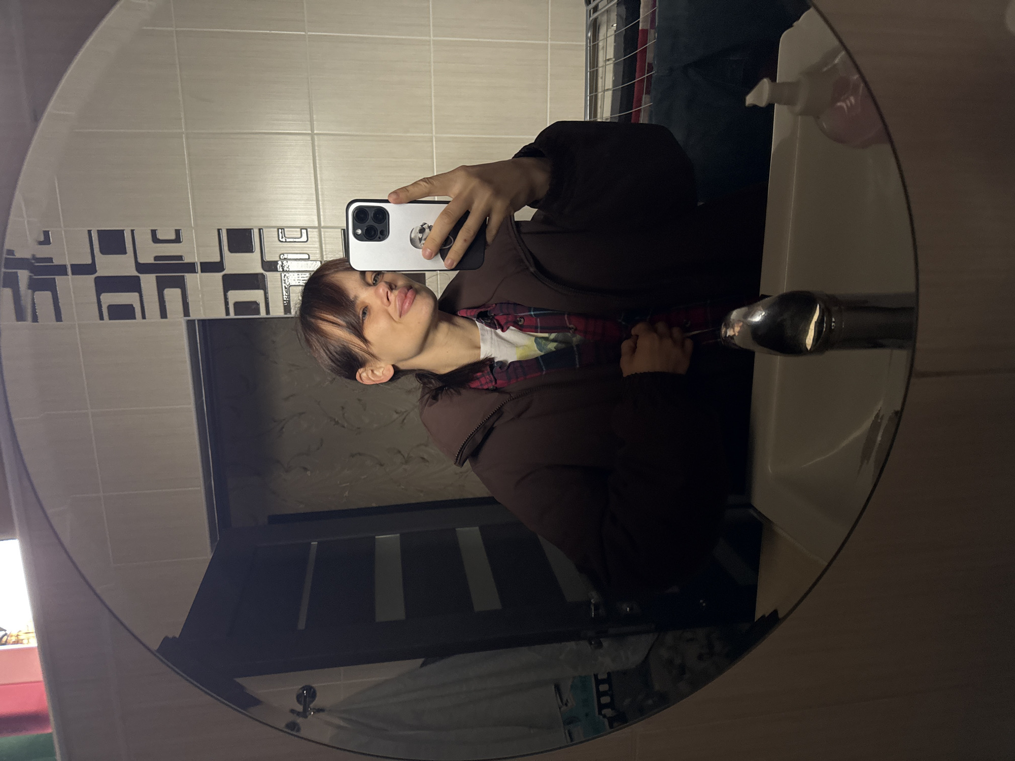 A young woman is taking a selfie in a bathroom mirror. She is holding a smartphone with a white case that has a skull design on the back. She is wearing a dark brown puffy jacket over a white t-shirt and a red and black plaid shirt. Her hair is brown with a blonde streak in her bangs.
The background includes beige and gray tiled walls with a black and gray decorative border. There is a white sink and faucet visible to the right of the woman, as well as a bottle of hand soap. A door with vertical black and white panels is visible in the reflection of the mirror. A partially visible washing machine is located in the background.
The location is a bathroom, likely inside a home or apartment. There is no indication the image was taken in Rostov-on-Don, Russia.