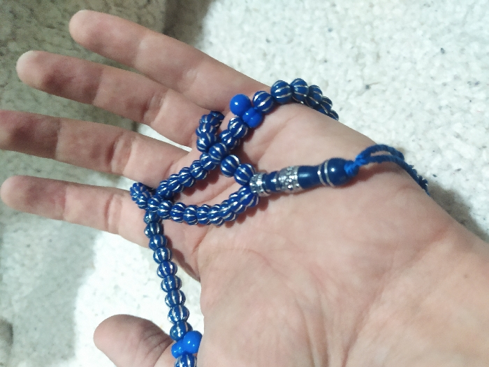 A human hand is depicted holding a string of blue prayer beads, likely within an indoor setting in the city of Unknown, Unknown. The hand, presented with the palm facing upwards, occupies the majority of the foreground, its fingers slightly curled around the beads. The prayer beads are primarily blue, consisting of spherical elements, many of which feature horizontal silver stripes. A larger, more ornate section of the beads, positioned towards the bottom center, includes solid blue beads interspersed with cylindrical silver-colored decorative components and terminates in a short blue cord. The background is a soft-focus, light-colored, textured surface, consistent with a carpet or similar textile. No other individuals or significant objects are visible in the scene.