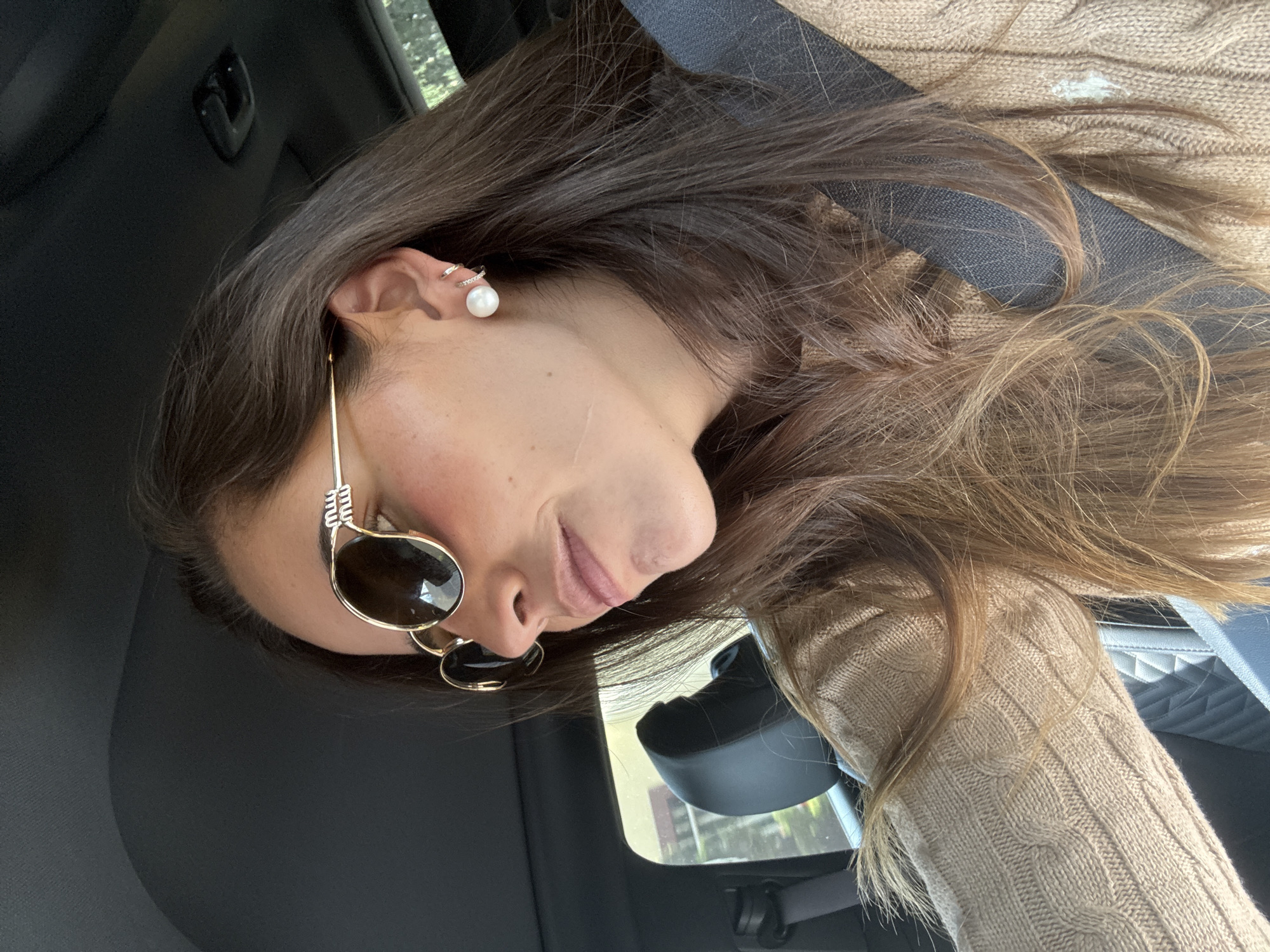 A close-up, side profile captures a person, likely a woman, sitting in a car during daylight hours. The individual has long, dark brown hair that flows over her shoulder and parts of her face. She is wearing gold-framed sunglasses with dark lenses, visible on the right side of her face. Her left ear is adorned with a pearl stud earring and a small, sparkling ear cuff on the upper cartilage. A light beige or tan cable-knit sweater is visible on her shoulder and arm.
The interior of the car features a dark-colored headliner and a visible seatbelt. A portion of a lighter-colored, possibly quilted, car seat or interior trim is also present. Through the car window, blurred green foliage and hints of buildings are discernible, suggesting an urban or suburban setting in San Bernardino Tlaxcalancingo, Mexico. Bright sunlight reflects off the sunglasses and illuminates the hair, indicating a sunny day. The person's lips are closed in a subtle, serene expression as she looks downwards and slightly away from the camera, conveying a sense of calm while in transit. There is no visible text in the image.