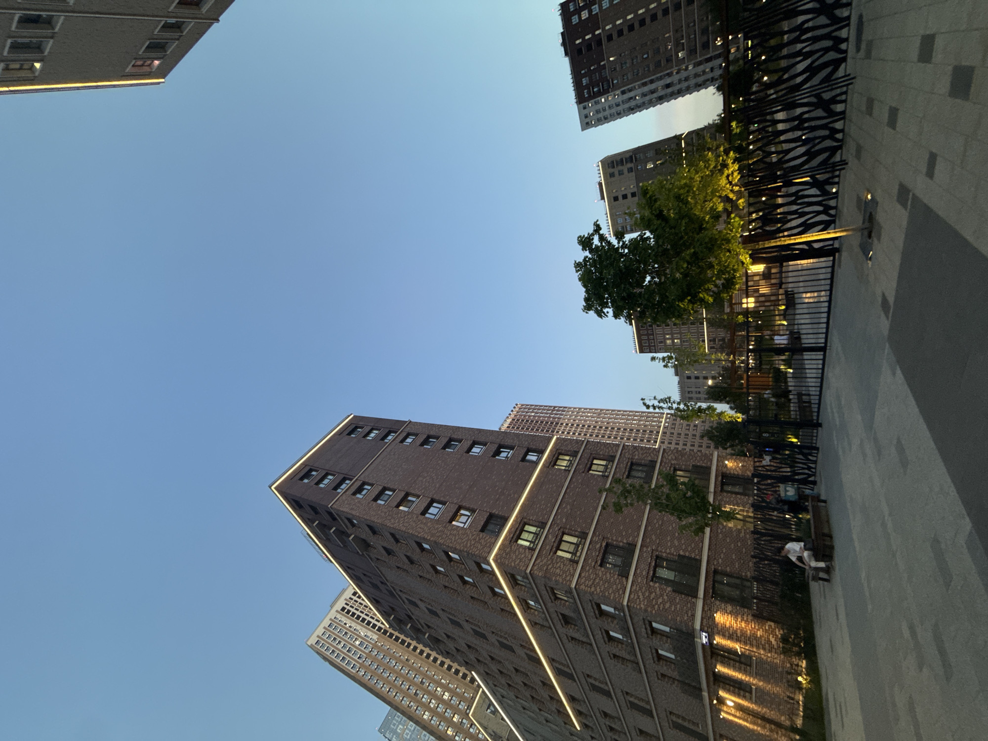 This low-angle shot depicts a modern urban environment in Moscow, Russia, based on the architectural style and signage (not visible but likely present). Several tall, multi-story buildings with brick facades and linear lighting accents are visible against a clear, light blue sky. A pedestrian area features decorative metal fencing, trees, and a paved walkway. A person dressed in light clothing is seated on a bench near the buildings. The perspective suggests the photograph was taken from street level, looking upward.