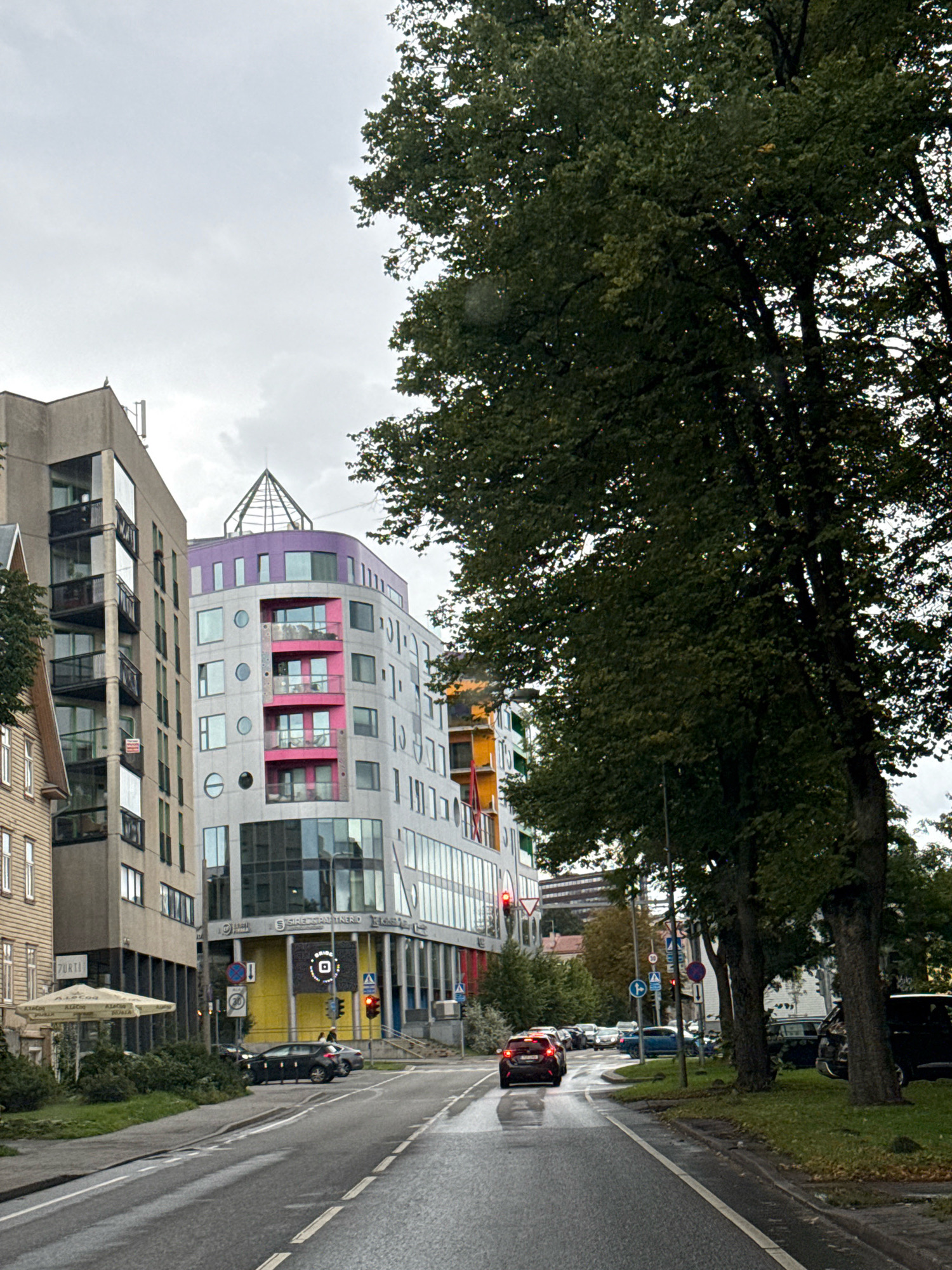 The image shows a view of a street in Tallinn, Estonia, with several modern buildings on the left and a line of trees on the right. The buildings are architecturally diverse, with a mix of glass, concrete, and colored panels. One building features a purple curved roof with a pyramidal structure on top and pink accents on its facade. Another building is taller and rectangular with balconies. The street is wet, suggesting it has recently rained, and there are several cars on the road. A red car is in the foreground, driving away from the camera. Traffic lights and street signs are visible. The sky is overcast.